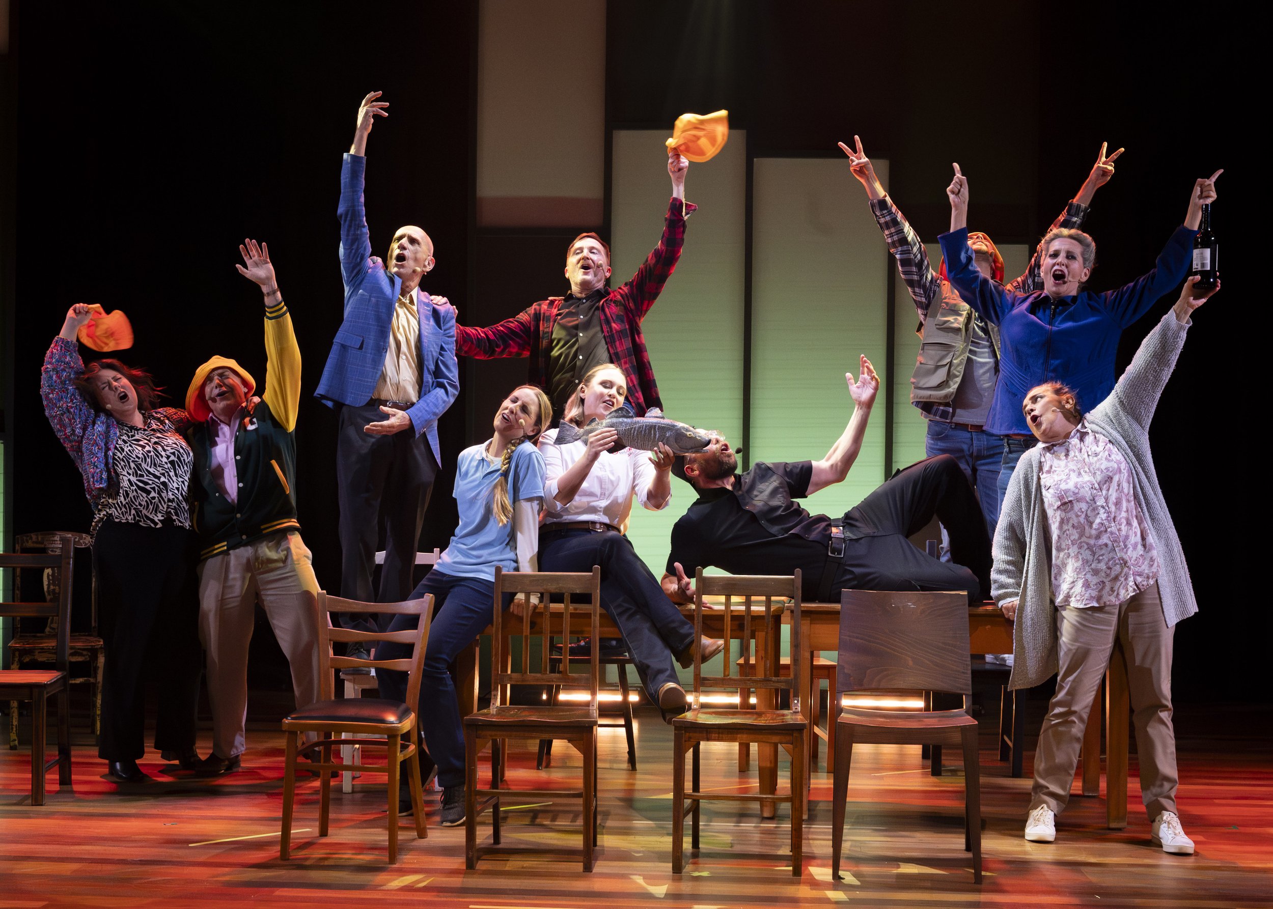 A lively stage scene with multiple people singing or acting, some sitting on chairs, some standing or kneeling, with animated gestures and expressions, holding hats and a bottle.