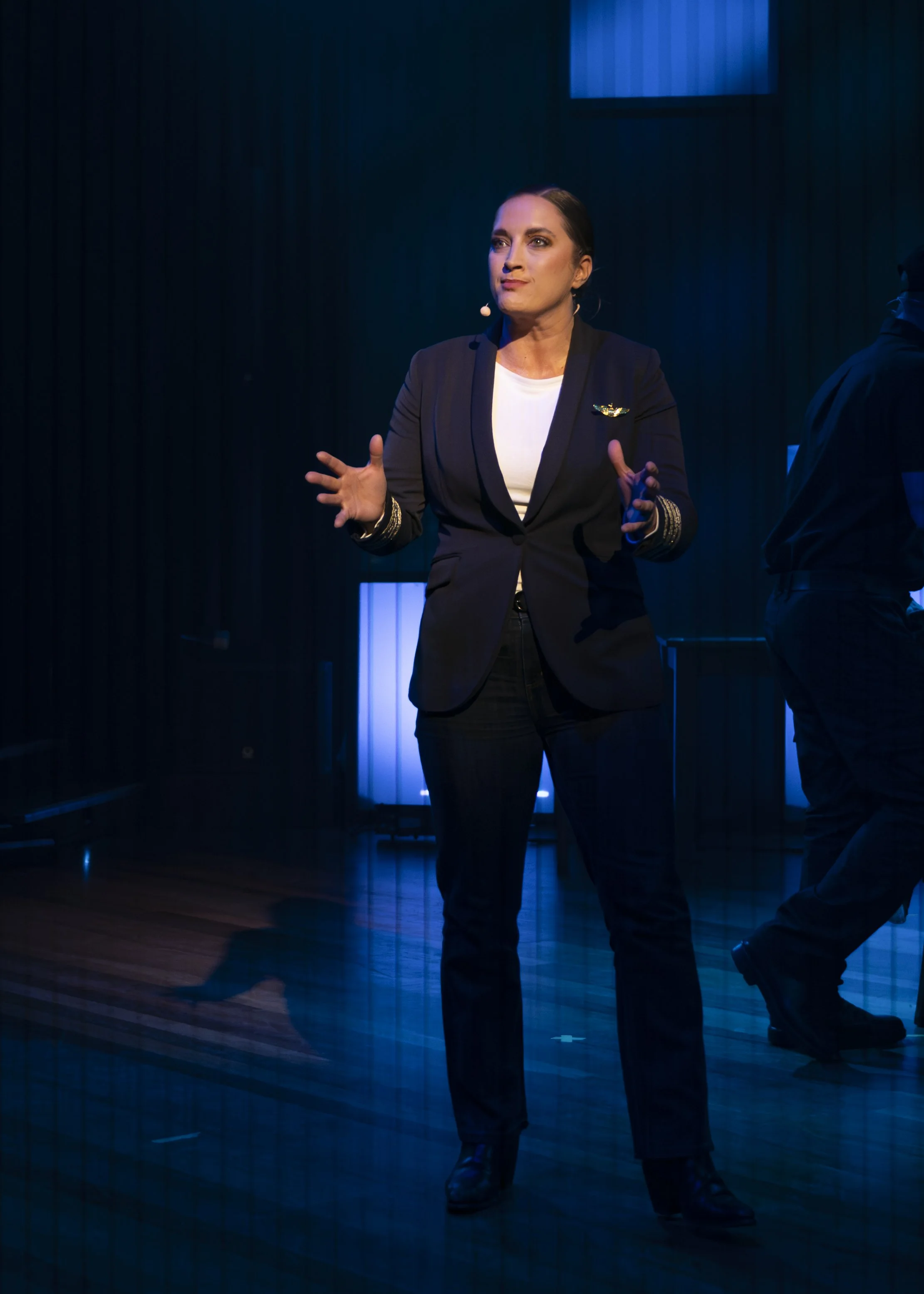 A woman in a black blazer and pants, white shirt, speaking on stage with dark curtains and blue lighting in the background.