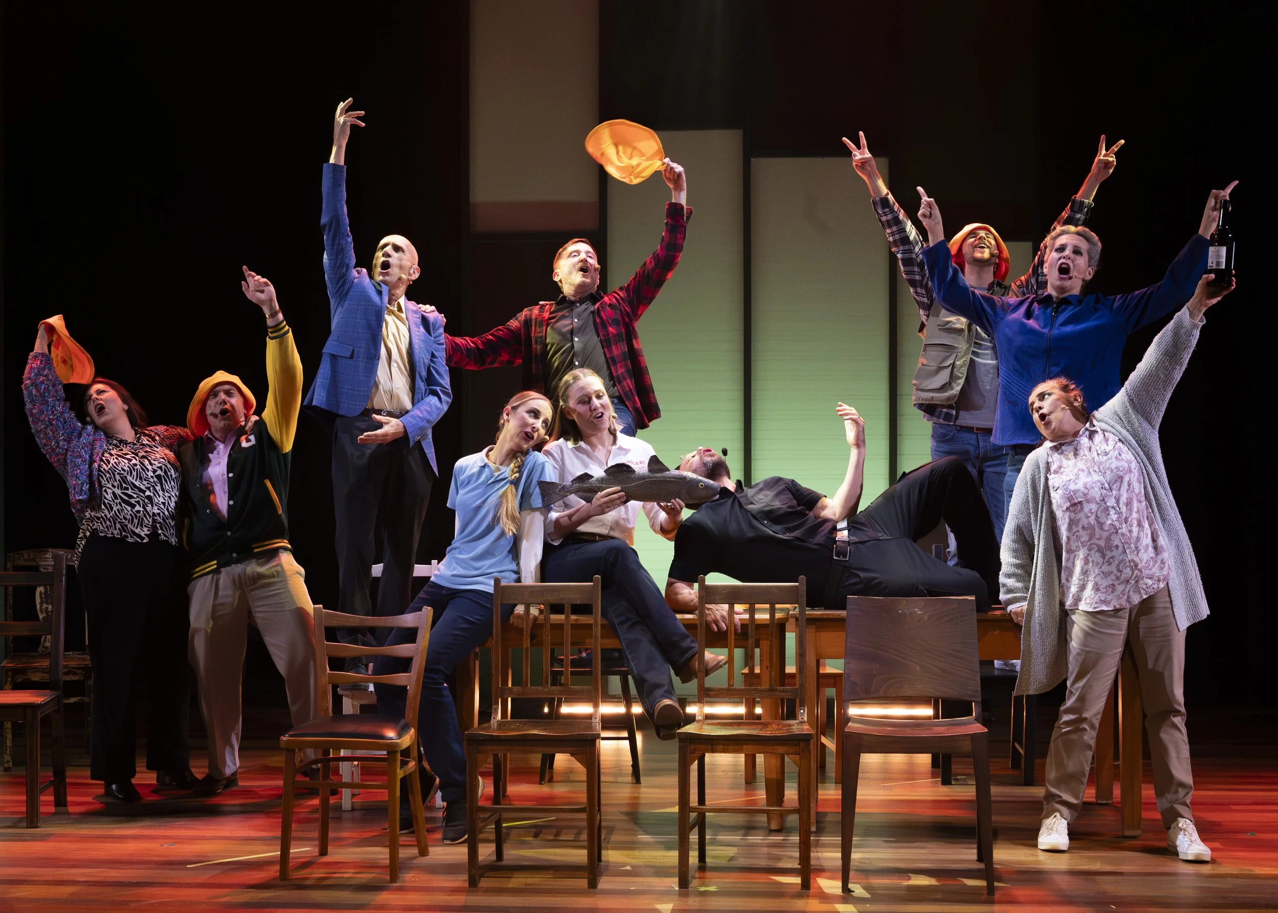 Actors performing on stage with exaggerated expressions, some raising arms, holding props like a fish, a bottle, and a hat, in a theatrical play.