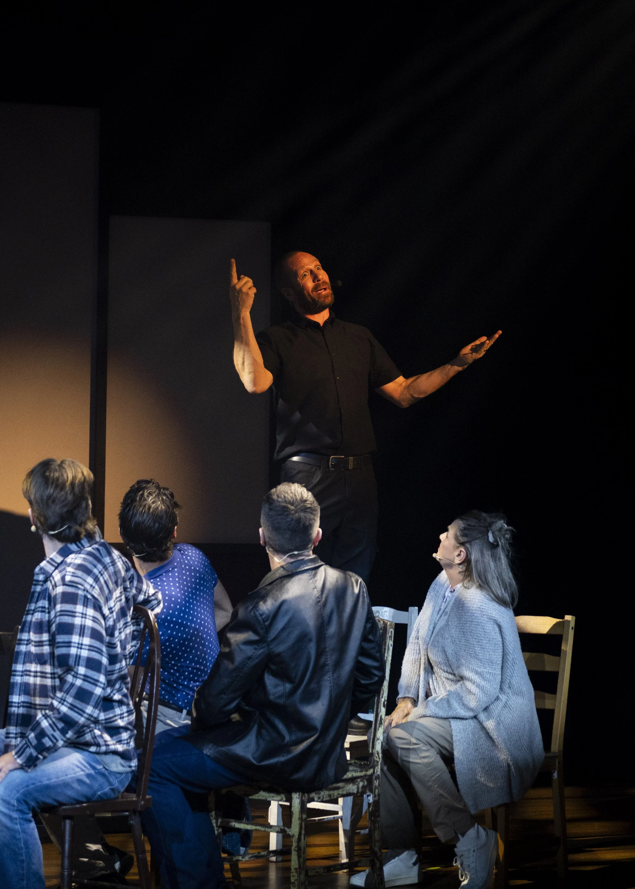 A man in black shirt giving a presentation to a seated audience in a dark room, gesturing with one finger raised.