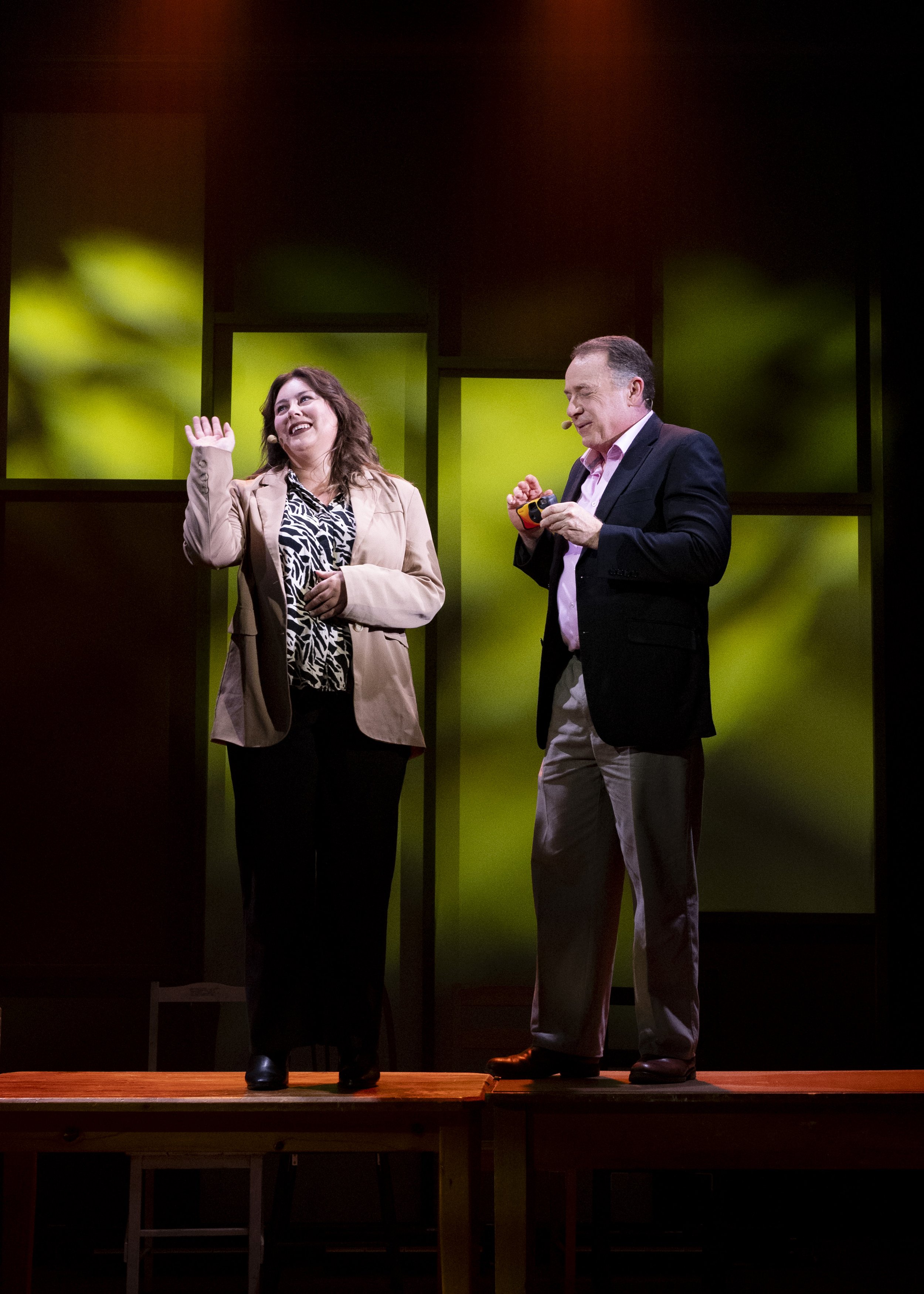 A woman and a man standing on a wooden stage, with the woman waving and smiling, and the man holding a small colorful object, both dressed in business casual attire, with a dark and green blurred background.