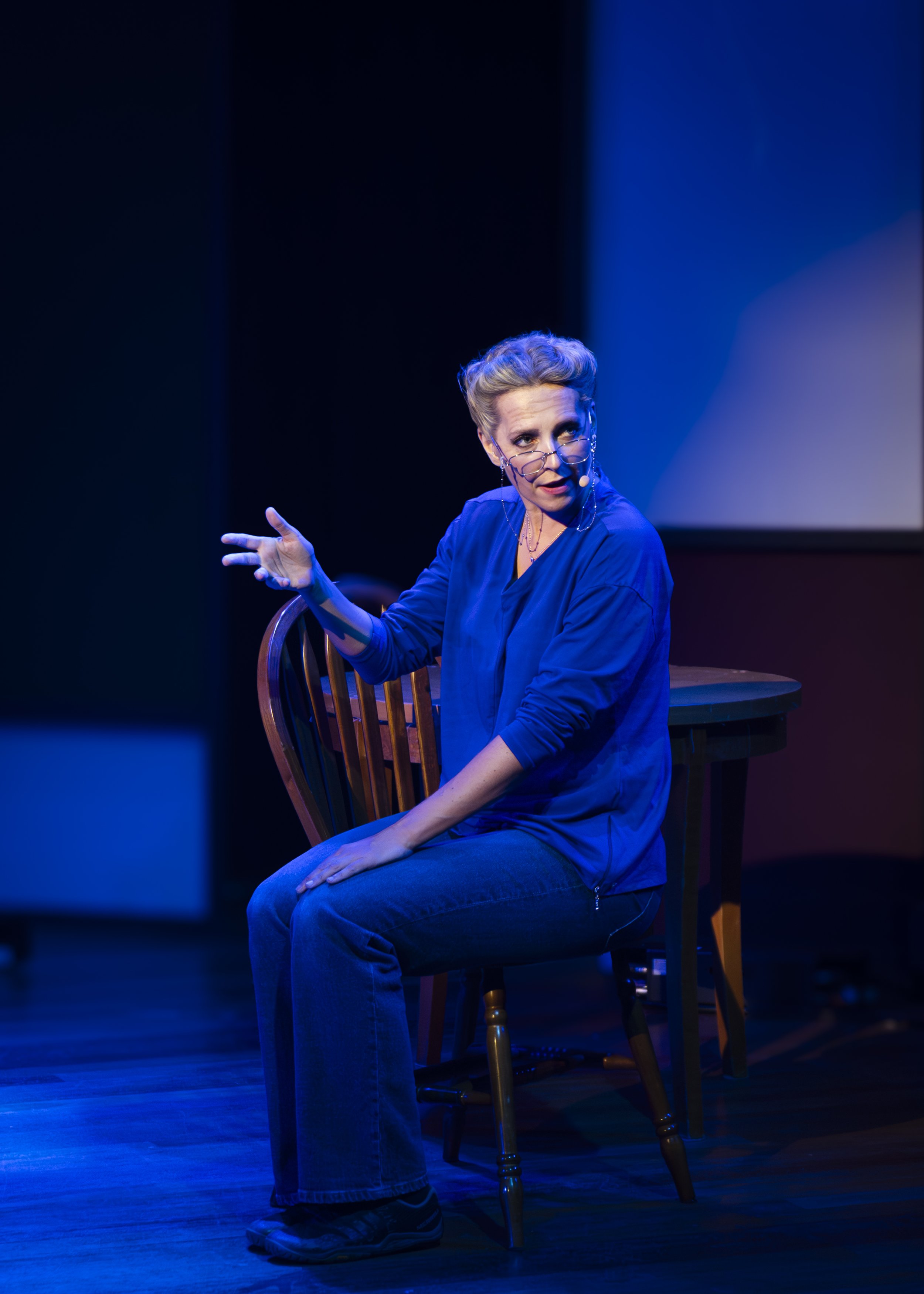 A woman with glasses and styled hair is performing on stage, seated on a wooden chair. She is wearing a blue jacket and jeans, gesturing with her left hand, under spotlight lighting.