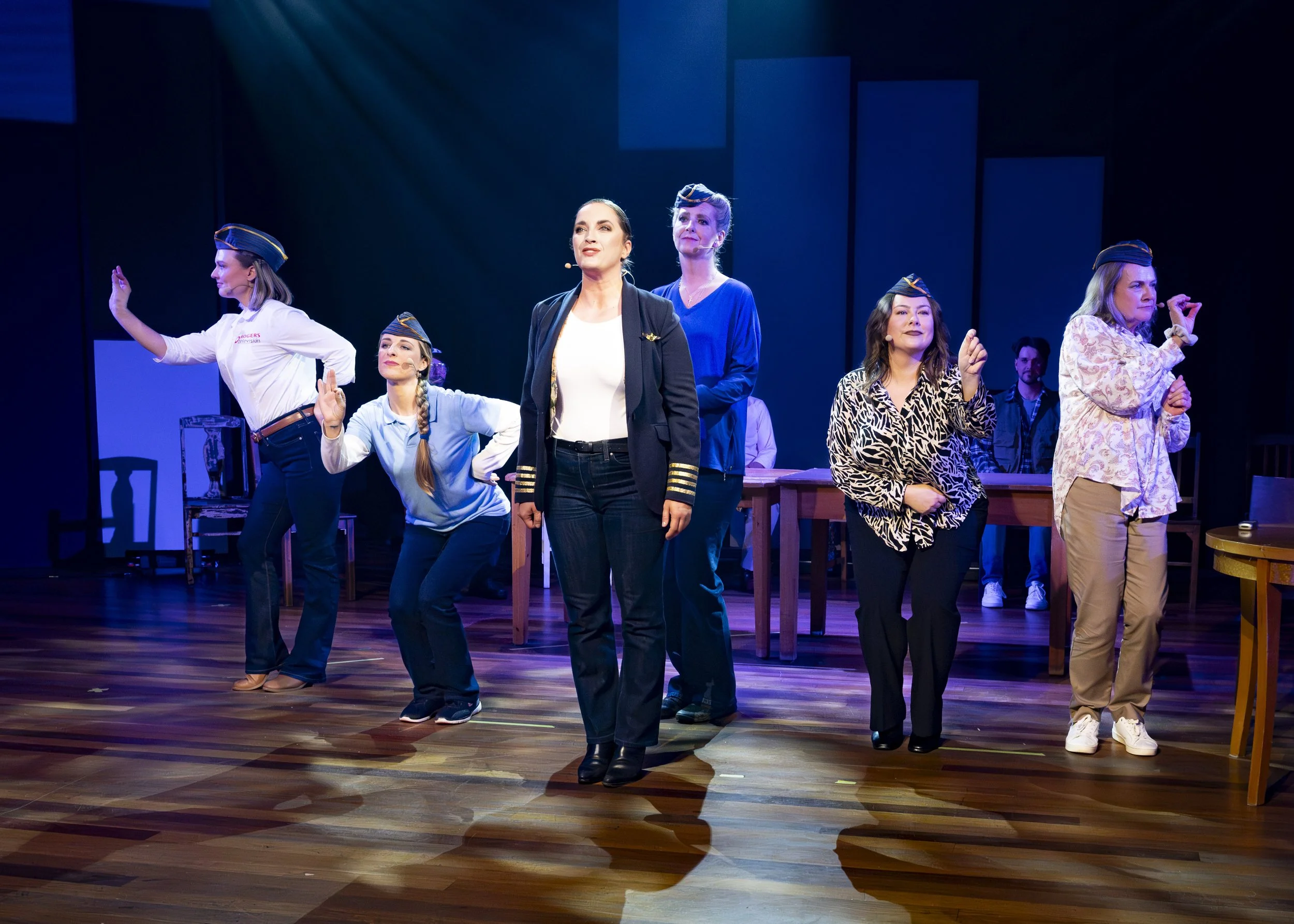 Theatrical performance on stage featuring six women, some in costumes and police hats, engaging in a scene with a table, chairs, and a dark background.