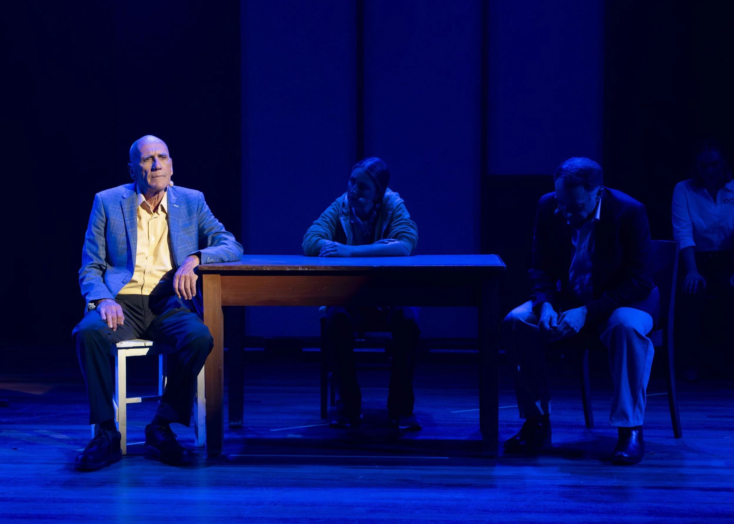 Three people sit at a wooden table on a stage under blue lighting, with a woman standing in the background.
