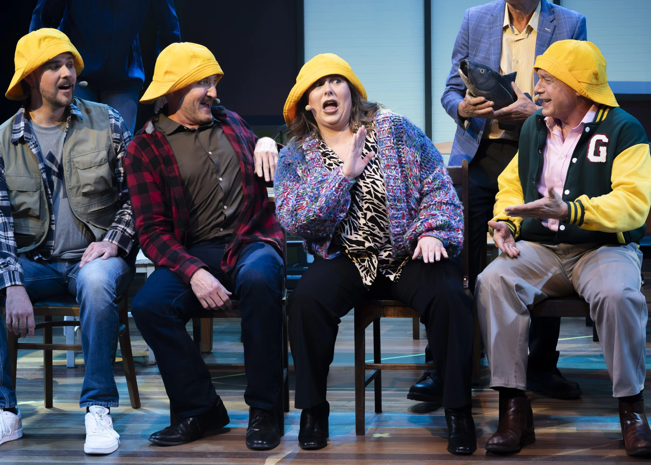 A group of six people on stage, with four sitting and two standing behind, performing a comedy sketch. The women and men are wearing colorful and casual attire, including yellow hats. One woman in the center appears surprised or scared, while others