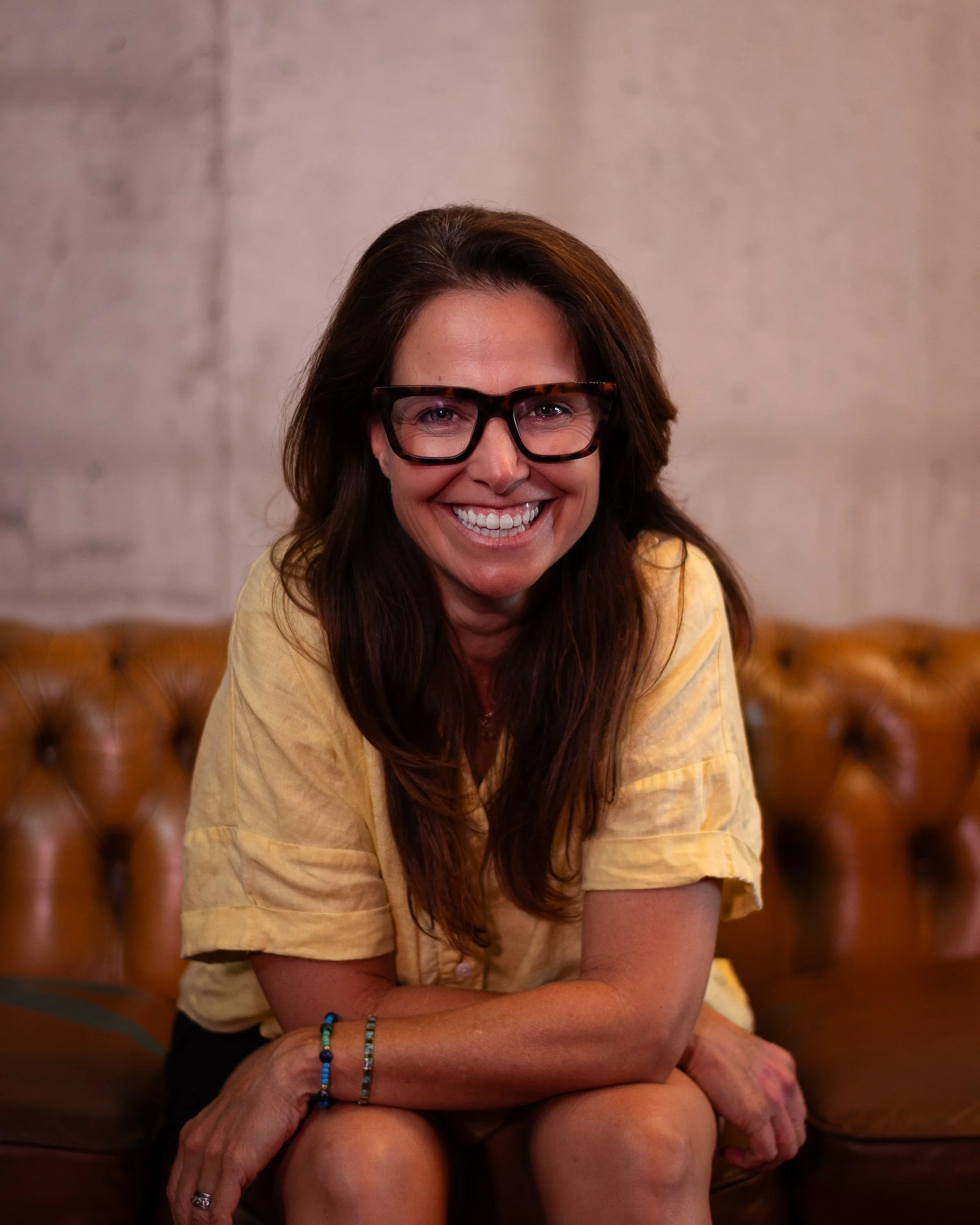 A woman with long brown hair, glasses, and a bright smile sitting on a brown leather couch with a concrete wall background.