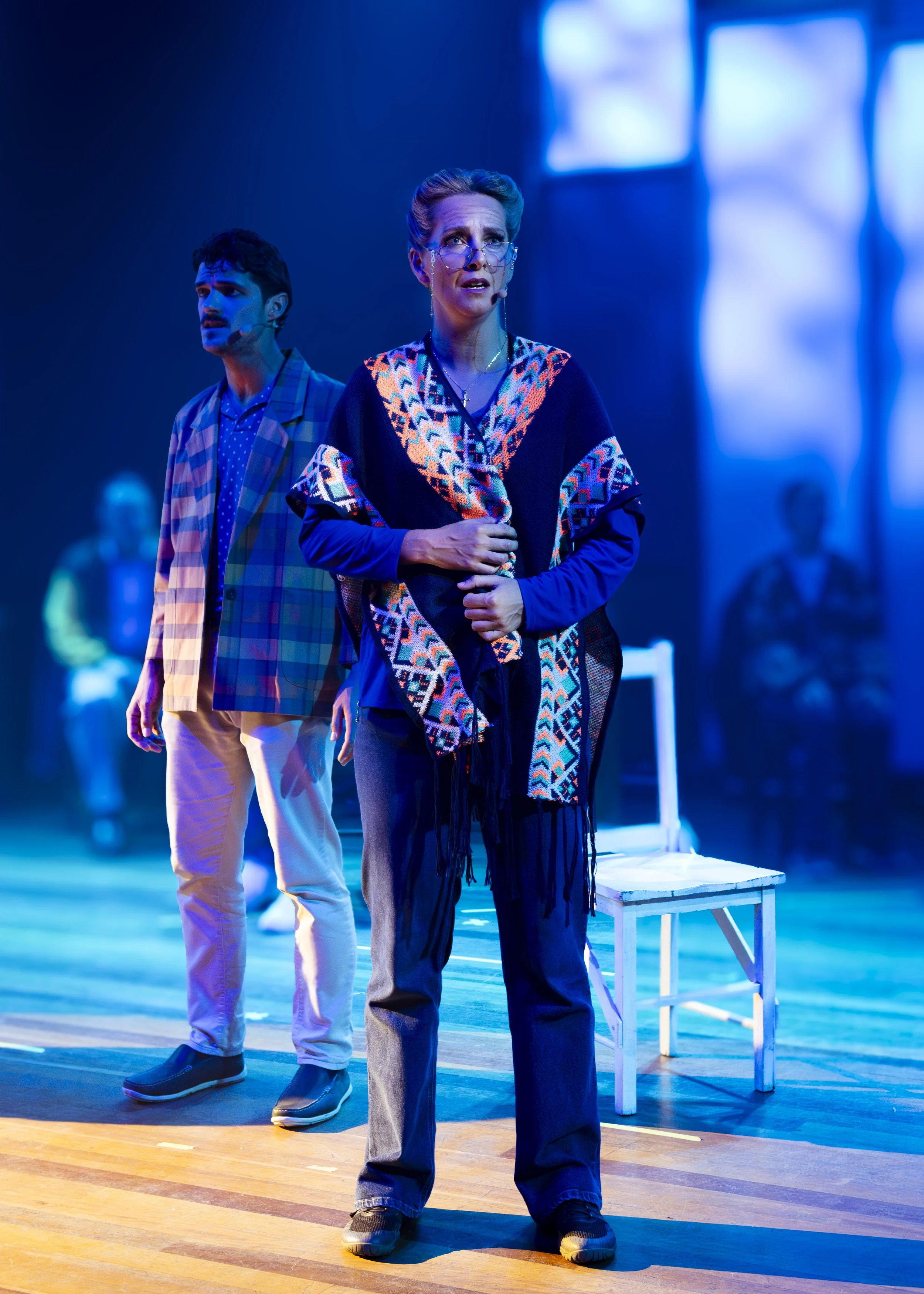 Two actors on stage during a theatrical performance, illuminated with blue lighting. The woman in the foreground is wearing glasses, a colorful shawl, and has a concerned expression. The man behind her is dressed in a checkered blazer and beige pants