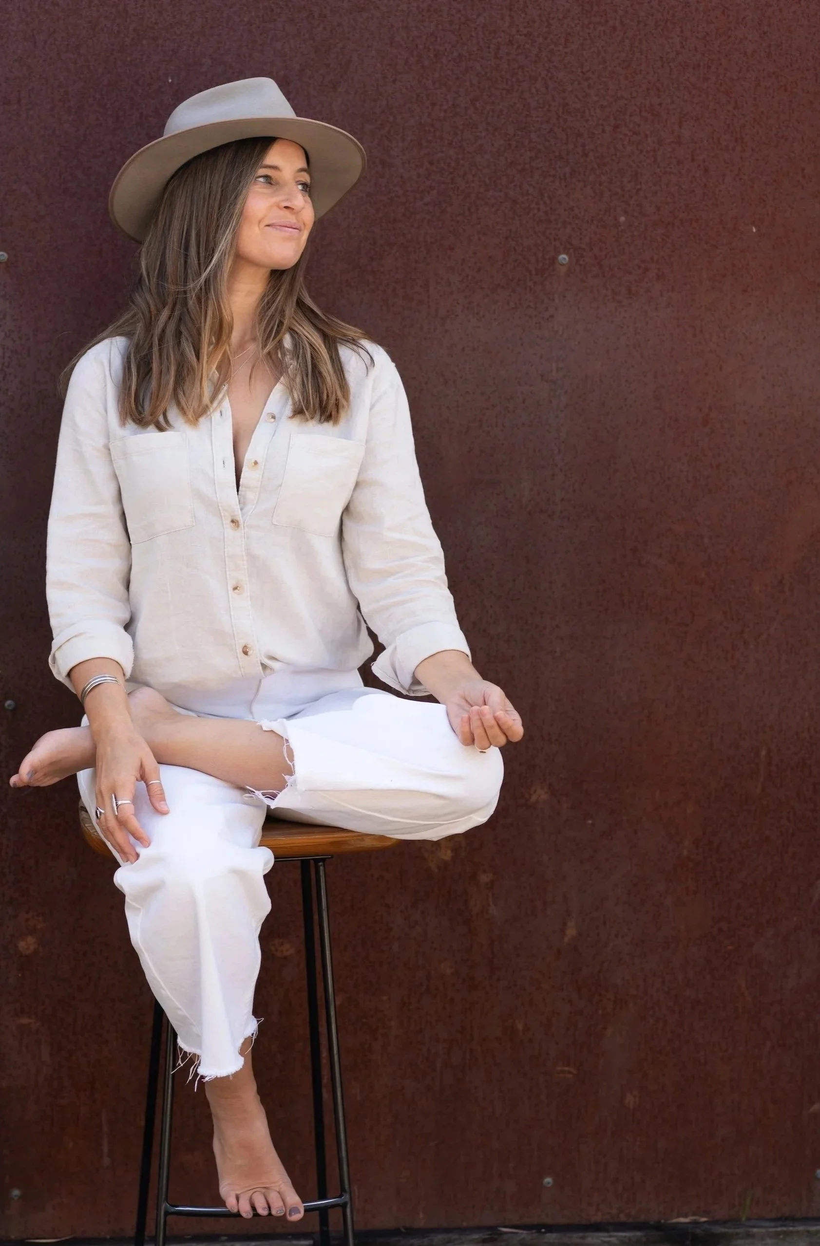 A woman with long hair wearing a beige hat, white shirt, and white pants practicing yoga or meditation while sitting on a stool against a brown textured wall.