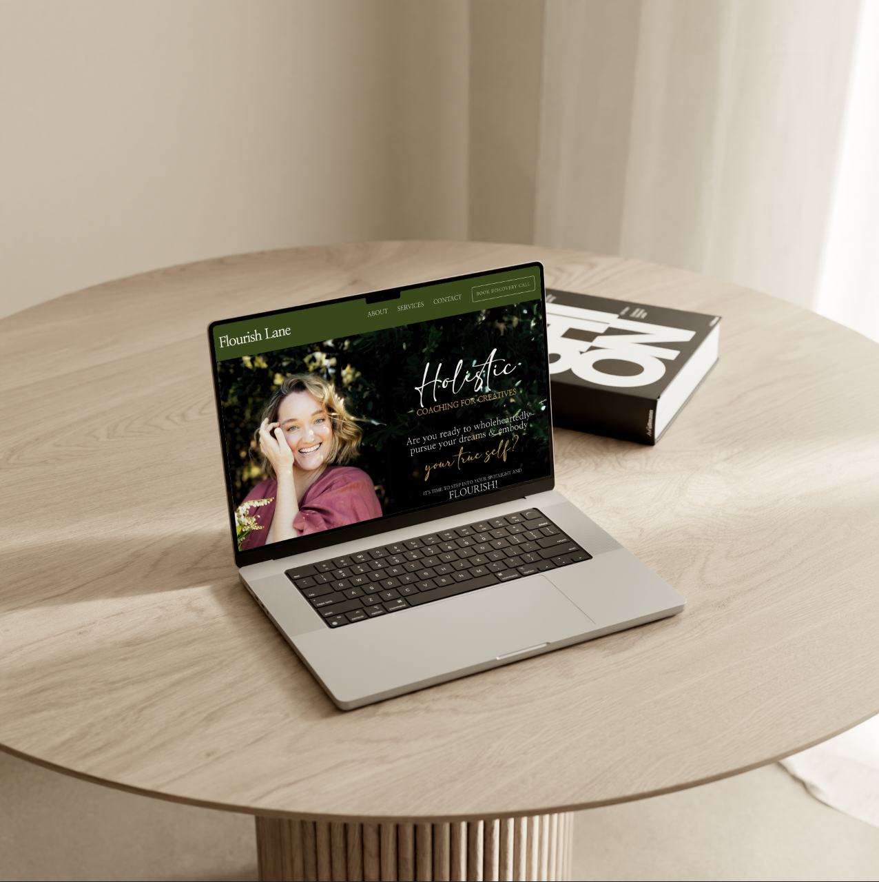 Laptop on a wooden table displaying a website for Flourish Lane, with a woman smiling, alongside a closed black and white book.