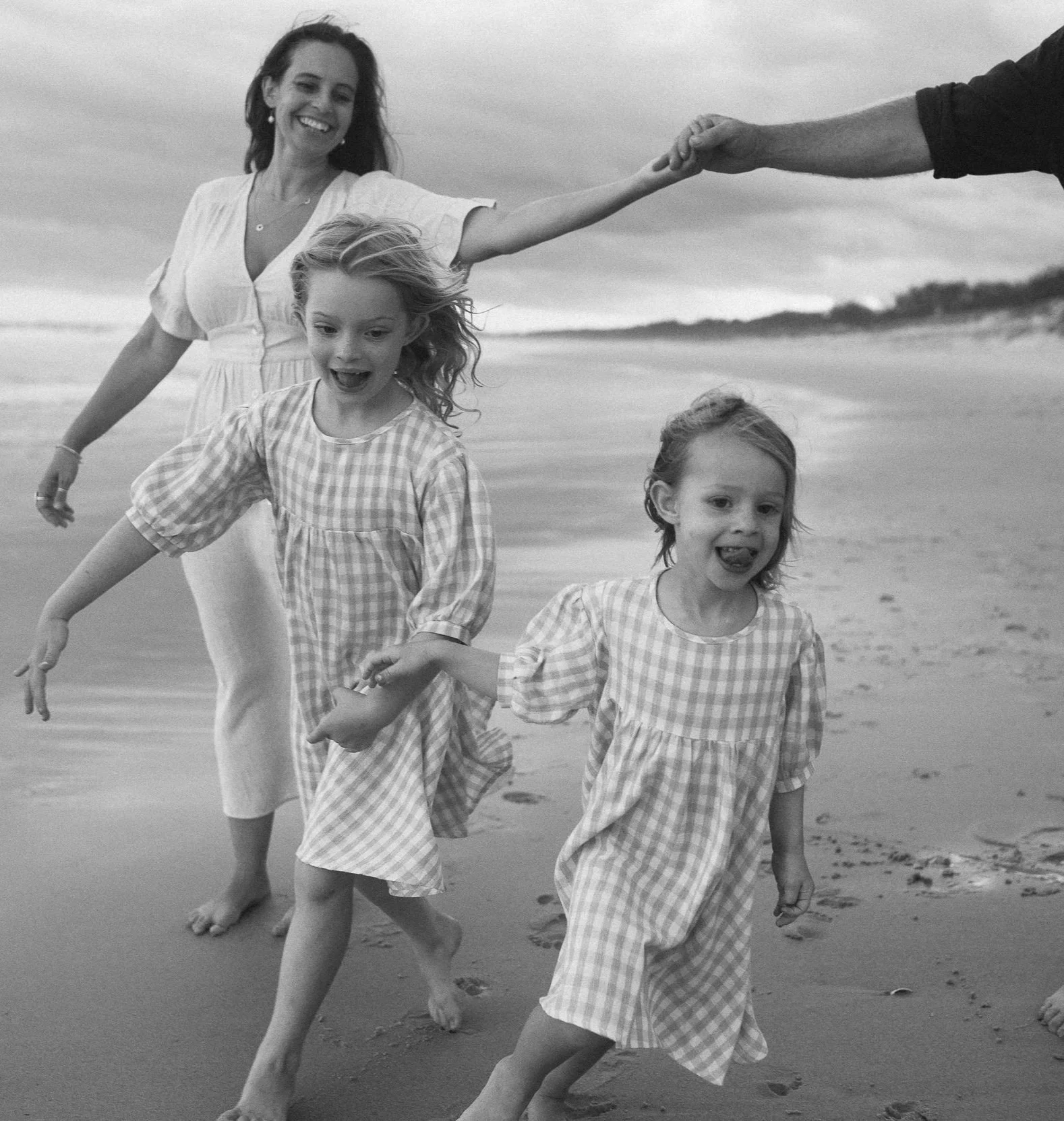 A woman and two young girls are running on a beach, holding hands, and enjoying a fun day. The image is in black and white.