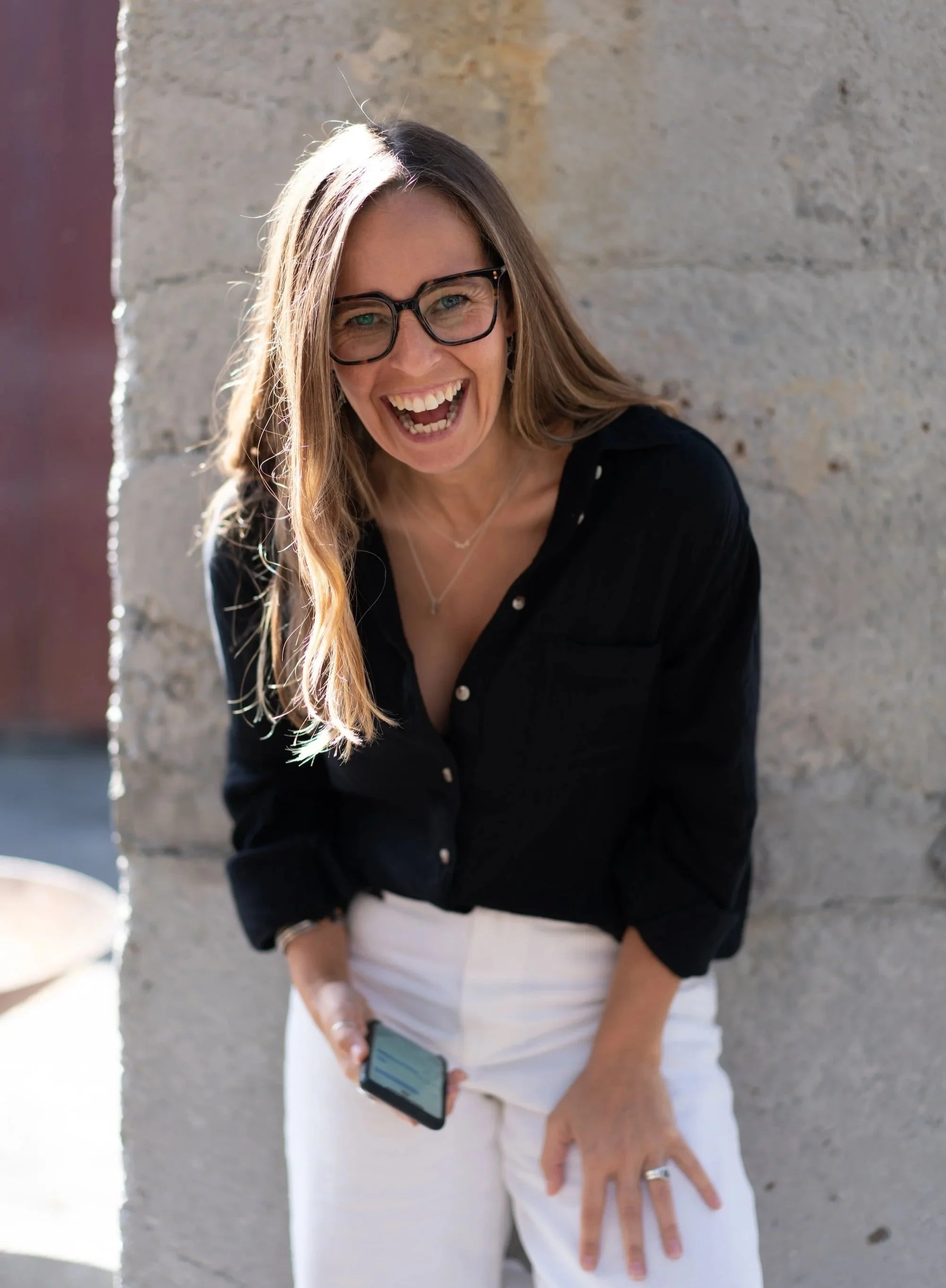 A woman with glasses and long hair laughing, holding a phone, standing next to a stone wall outside.