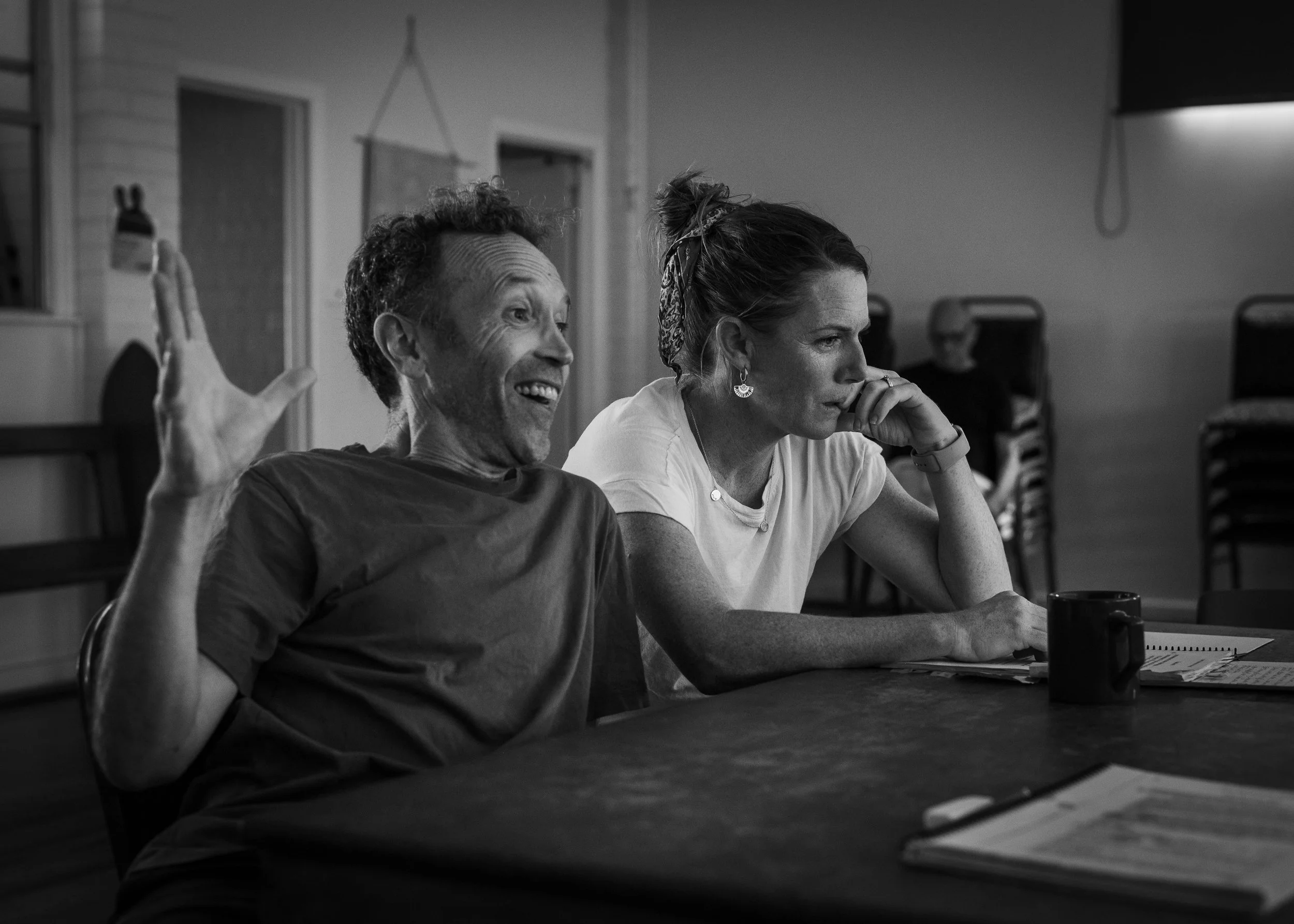 Two women sitting at a table in a room, one smiling and gesturing with her hand, the other resting her chin on her hand and listening thoughtfully, with notebooks and a mug in front of them.
