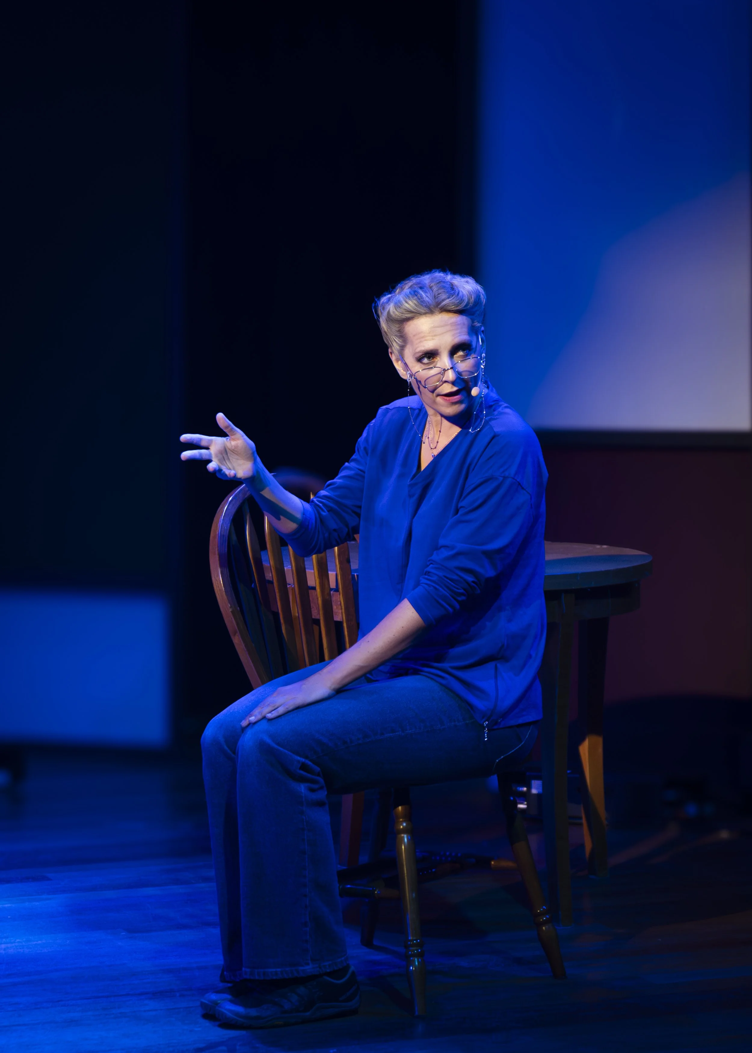 A woman with glasses and blonde hair, dressed in a blue top and jeans, sitting on a wooden chair on a stage, gesturing with her hand while speaking, illuminated by stage lighting.