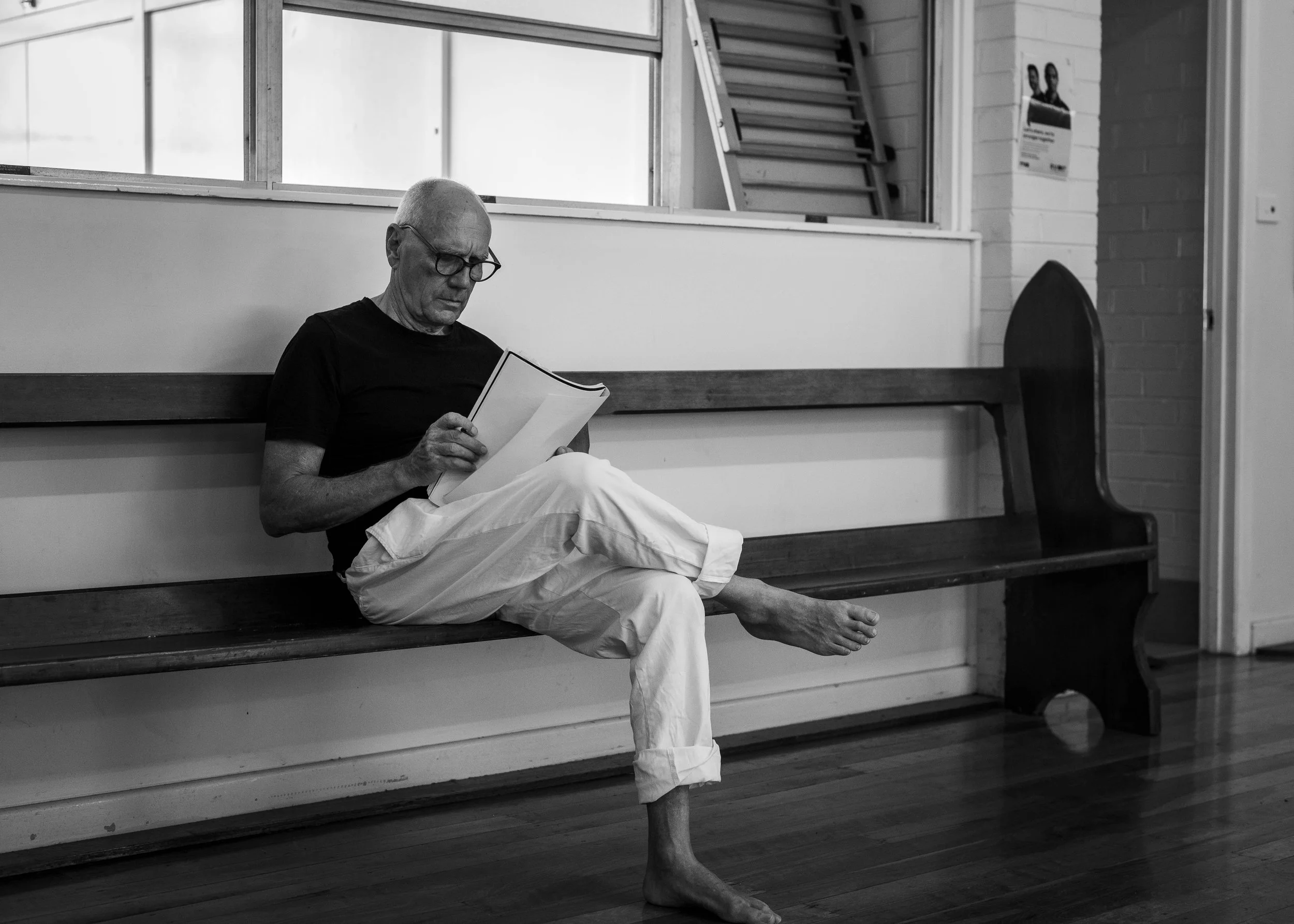 An elderly man with glasses wearing a black shirt and white pants sitting barefoot on a wooden bench, reading a book, in a room with large windows and a brick wall.