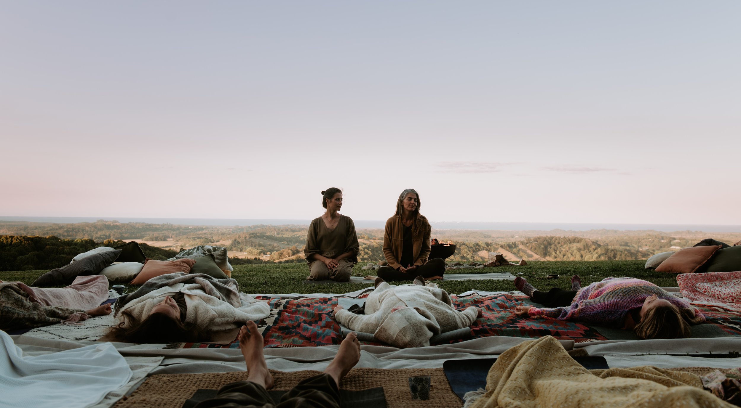 People lying on blankets and pillows on the grass during sunset, with two women sitting and meditating or relaxing in the background overlooking a landscape.