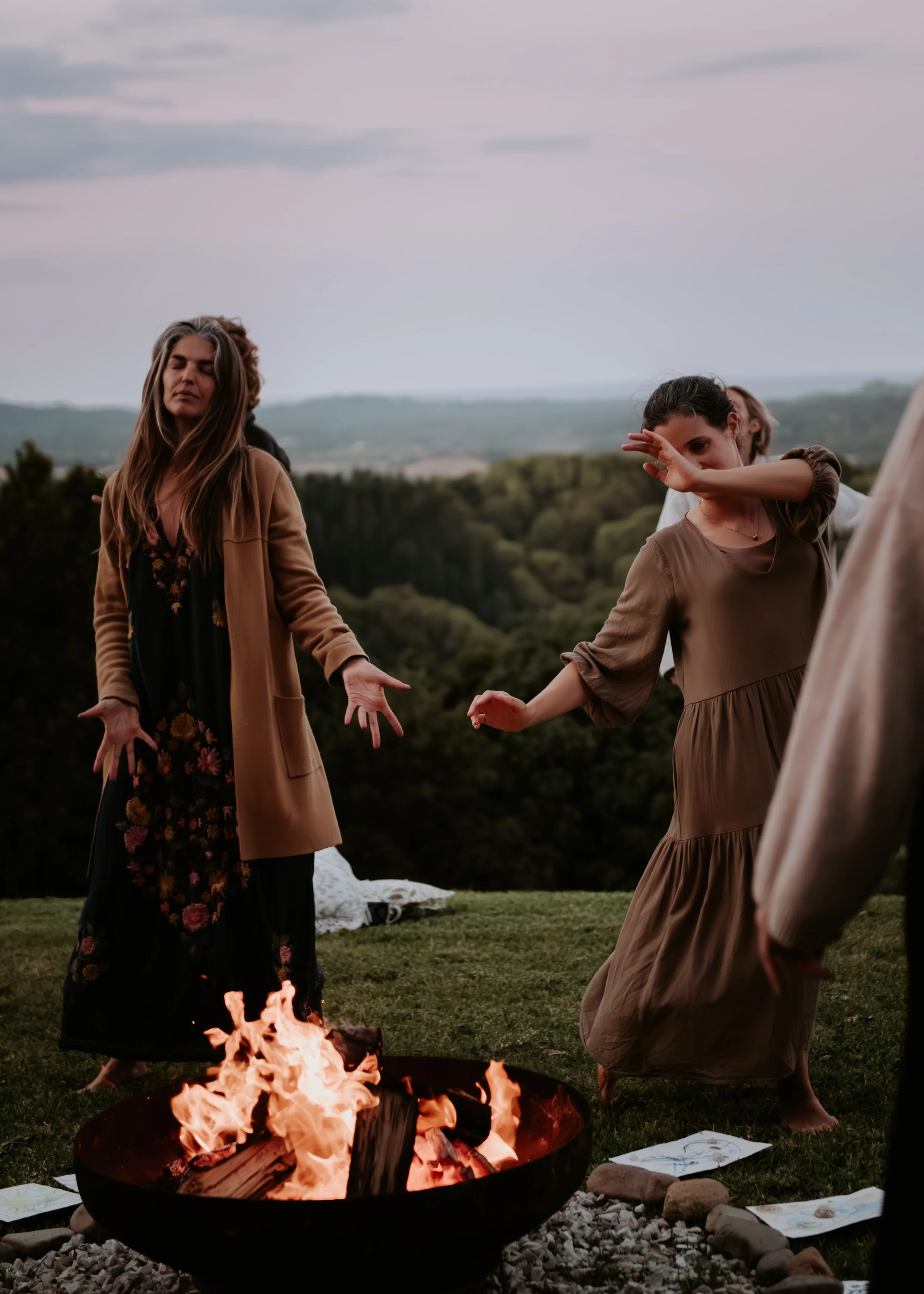 People standing around a fire outdoors, some with their eyes closed or covering their faces, with a scenic landscape in the background during dusk or evening.