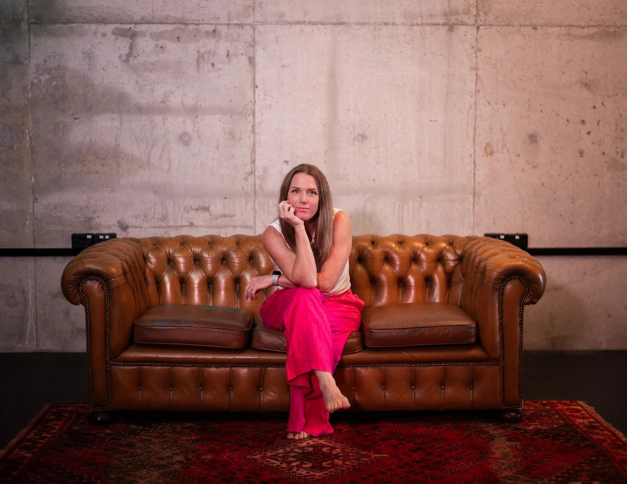 A woman with long brown hair sits on a brown leather tufted sofa, resting her chin on her hand, wearing a beige sleeveless top and bright pink pants. She is in front of a plain concrete wall with visible texture and lines, with a patterned rug undern