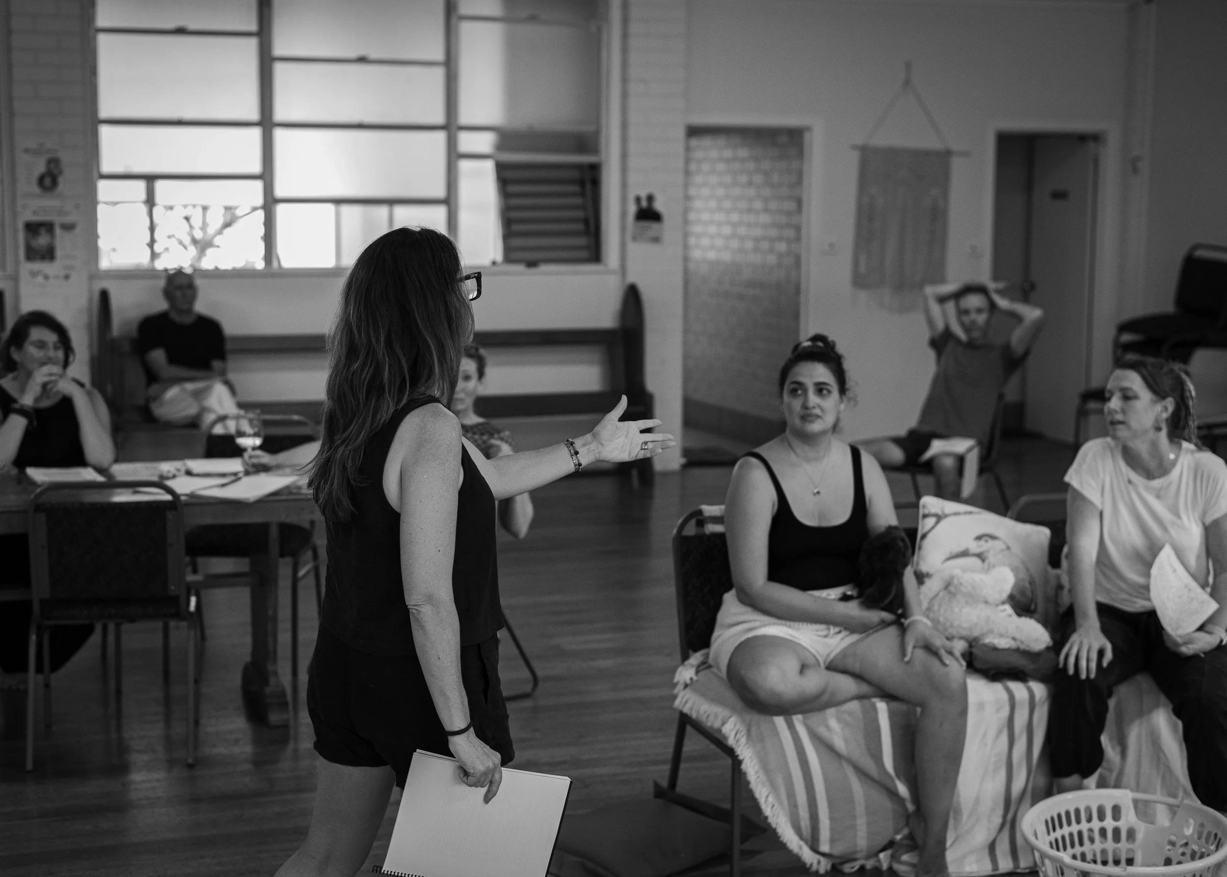 A woman with glasses and long hair is speaking or presenting to a group of people in a room. The group includes women sitting on a sofa and chairs, some with notebooks or papers, and a man sitting in the background. The room has large windows and var