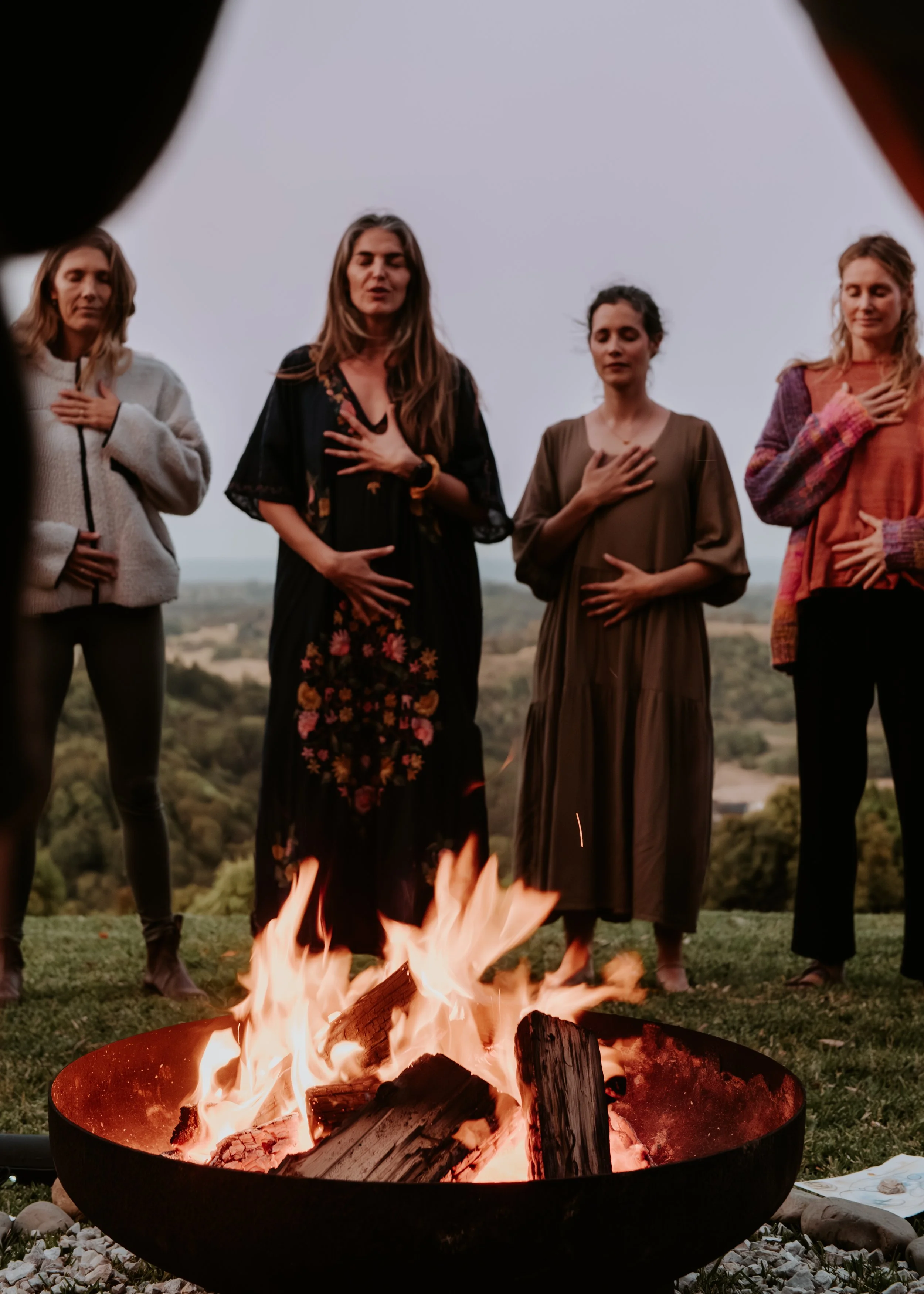 Four women standing with their hands over their hearts around a fire pit outdoors, with a scenic landscape in the background.