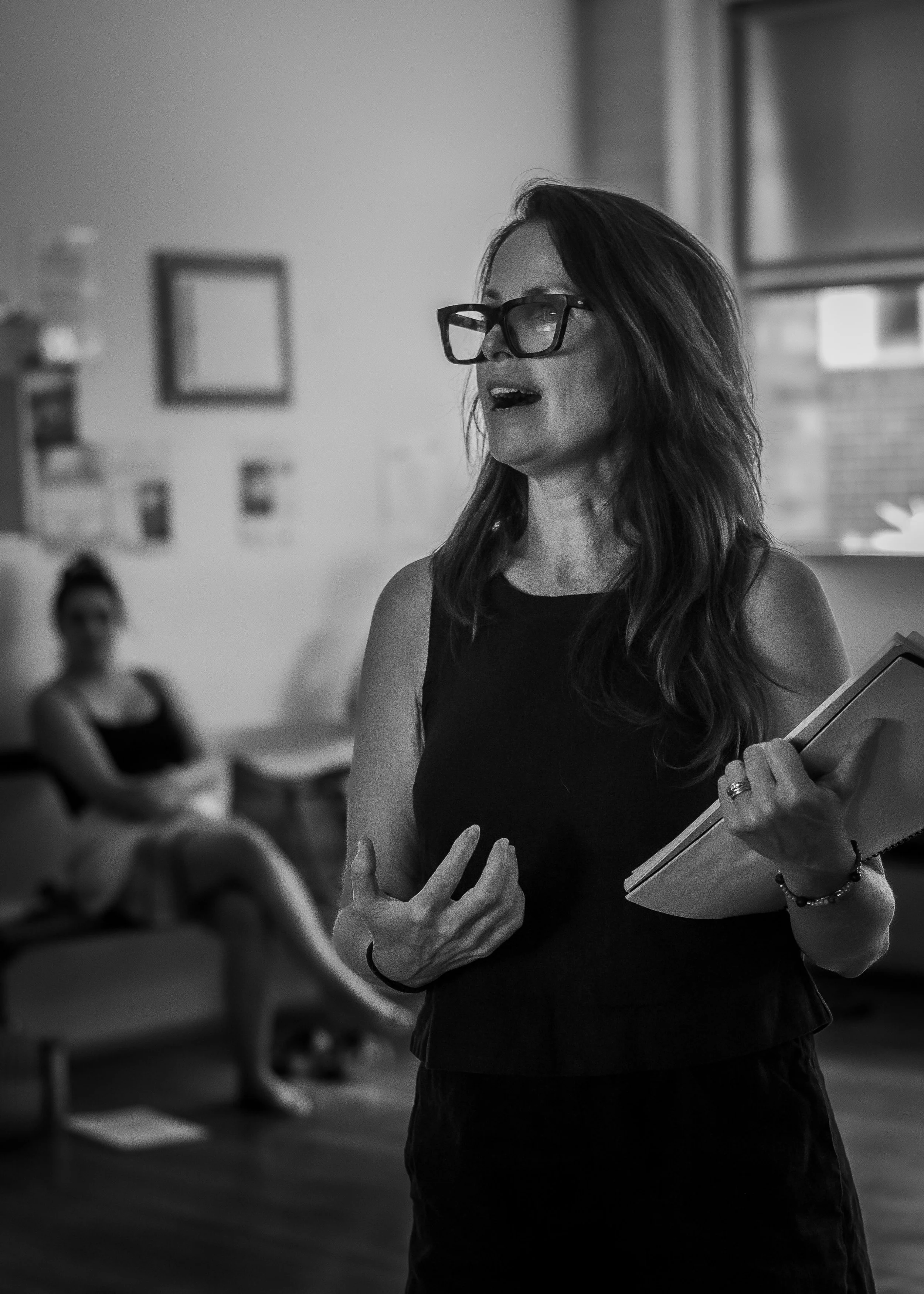 A woman with glasses speaking or giving a presentation in a room, holding a notebook, with another woman sitting in the background.