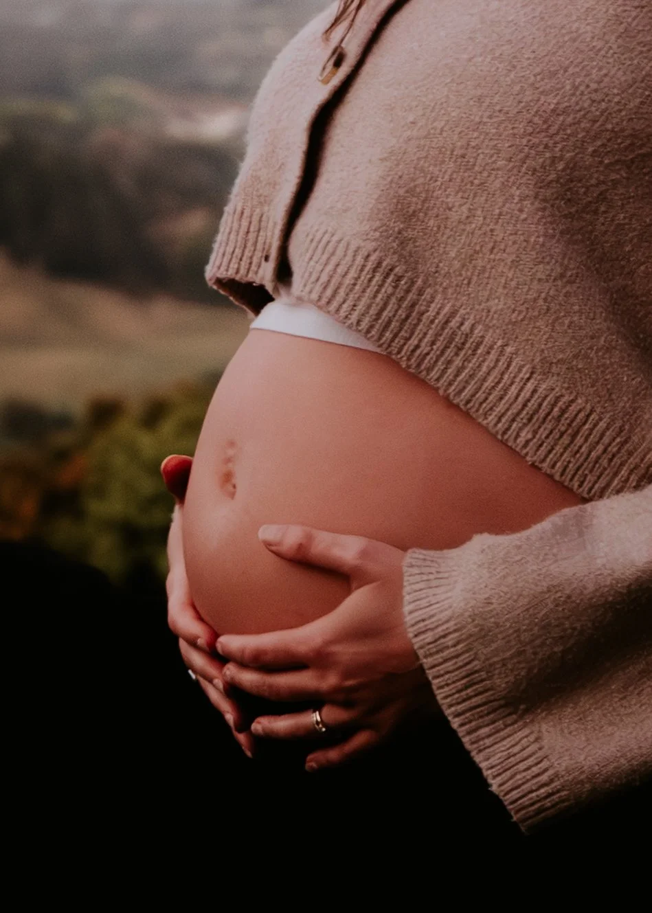 Close-up of a pregnant woman's belly with hands gently resting on it, wearing a beige sweater with a portion lifted to reveal the belly.