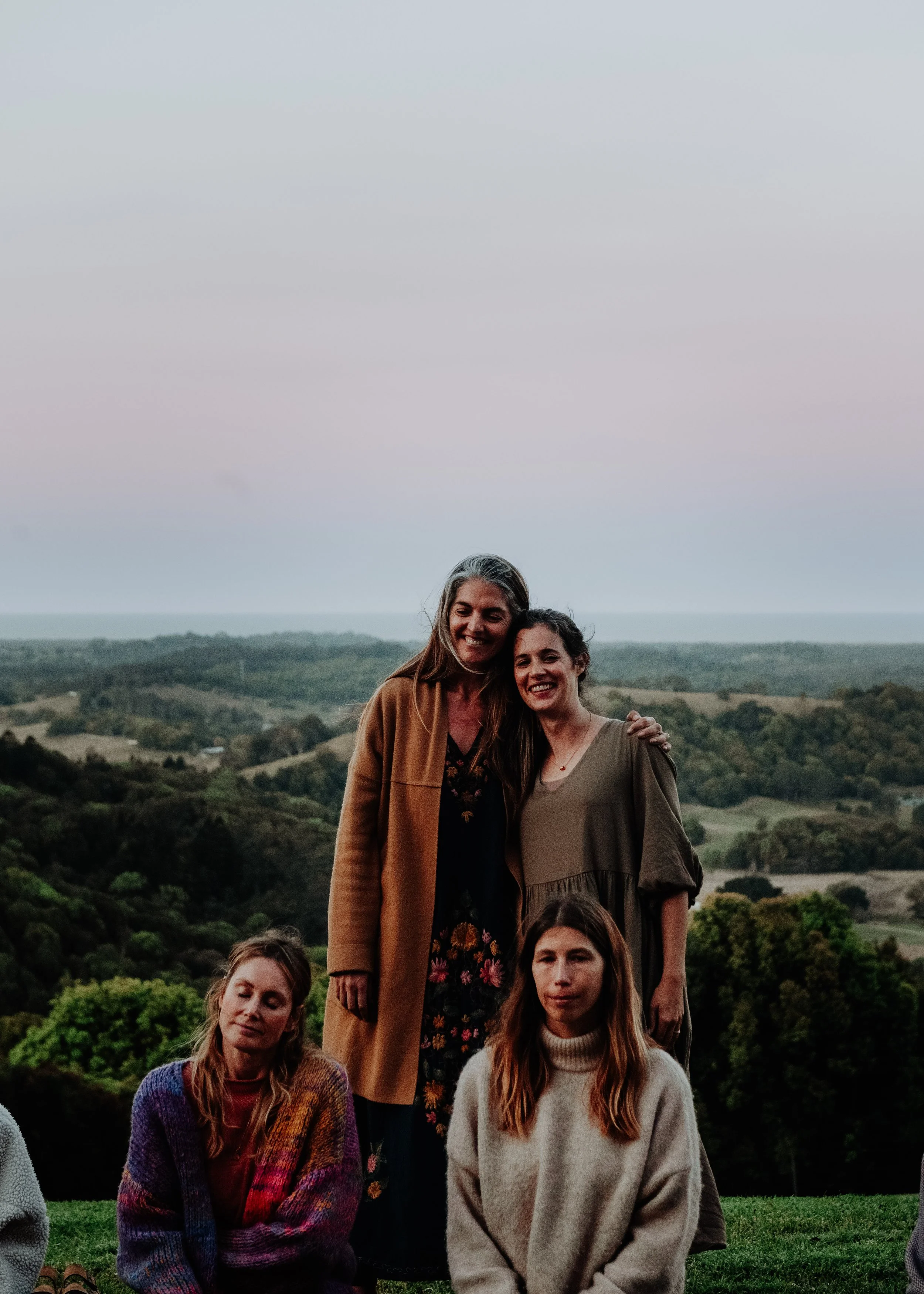 Two women standing outdoors on a hilltop, smiling, with a scenic landscape of rolling hills and trees in the background. Two other women are sitting in front of them, one with a colorful sweater and the other with a beige sweater.