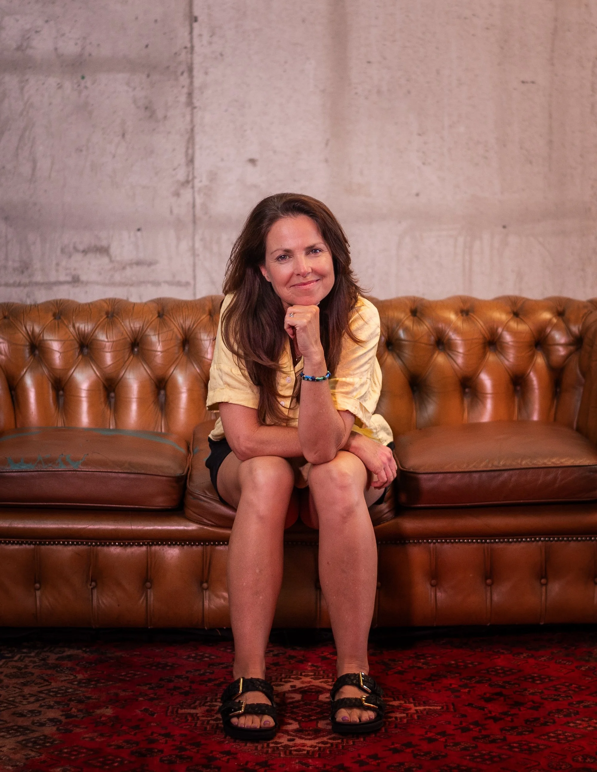 A woman with long brown hair sitting on a worn brown leather tufted sofa, wearing a yellow shirt, black shorts, and black sandals, smiling with her chin resting on her hand, in front of a concrete wall.