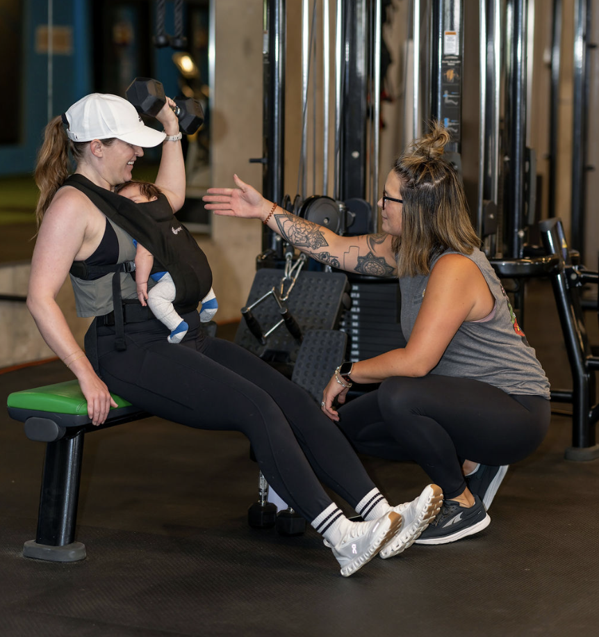 A woman lifting a barbell with weights in a gym, seen from behind, wearing a white sports bra and black leggings.