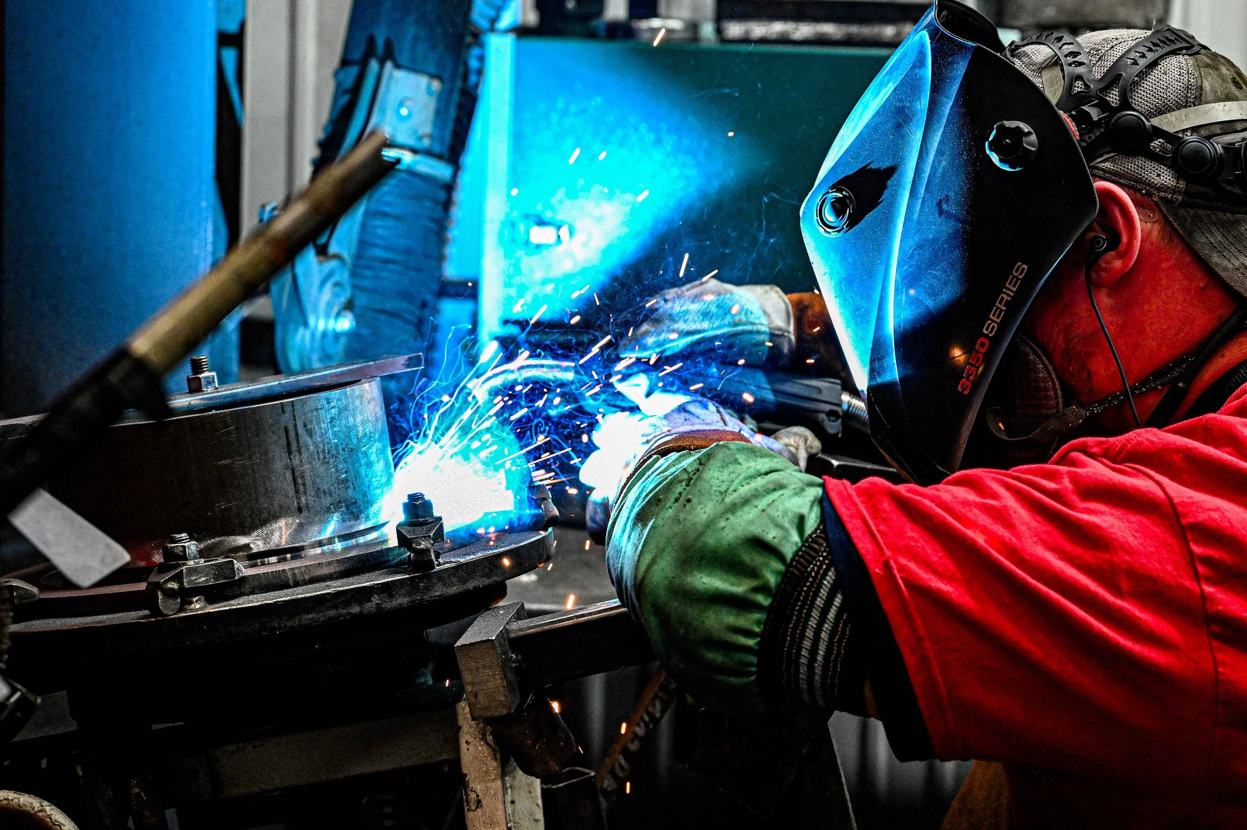 A person welding metal in a workshop, wearing protective gear including a welding helmet with a blue reflective visor and gloves, sparks flying as they work on a metal piece.