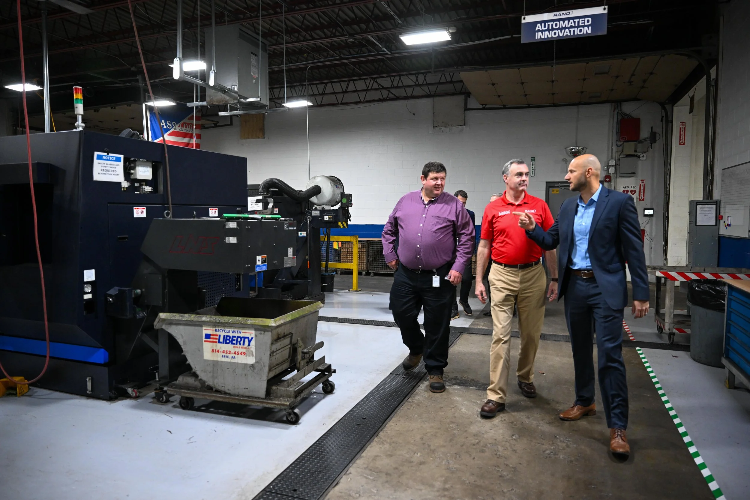 Dave Malone, Dan McKenrick, Peter Tur walking in an industrial or manufacturing facility. They appear to be having a discussion. The area contains machinery and equipment, with signs and safety devices visible.