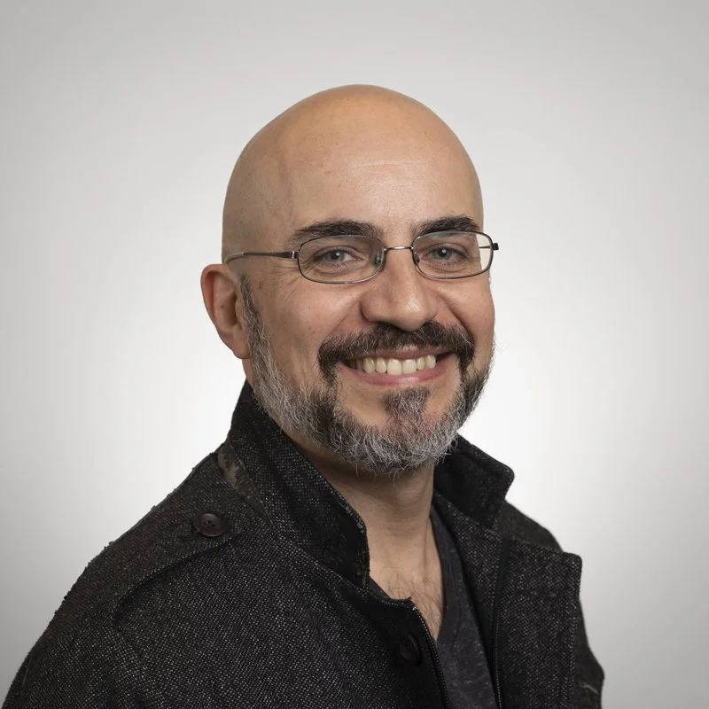 Headshot of a smiling bald man with glasses, a beard, wearing a black textured jacket, against a plain light gray background.