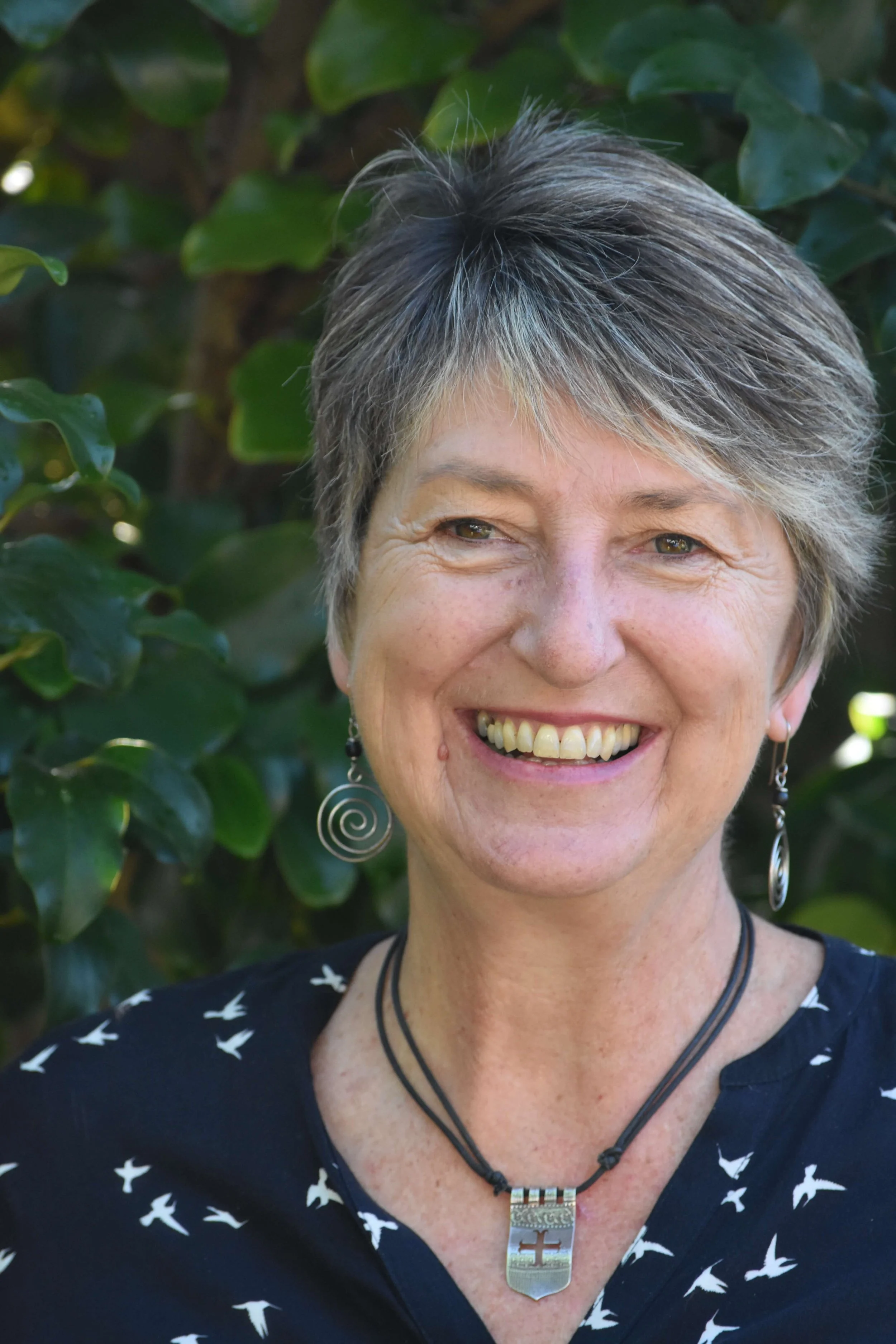 A smiling woman with short gray hair, wearing earrings, a black necklace, a pendant, and a black top with white bird prints, standing in front of green leafy bushes.