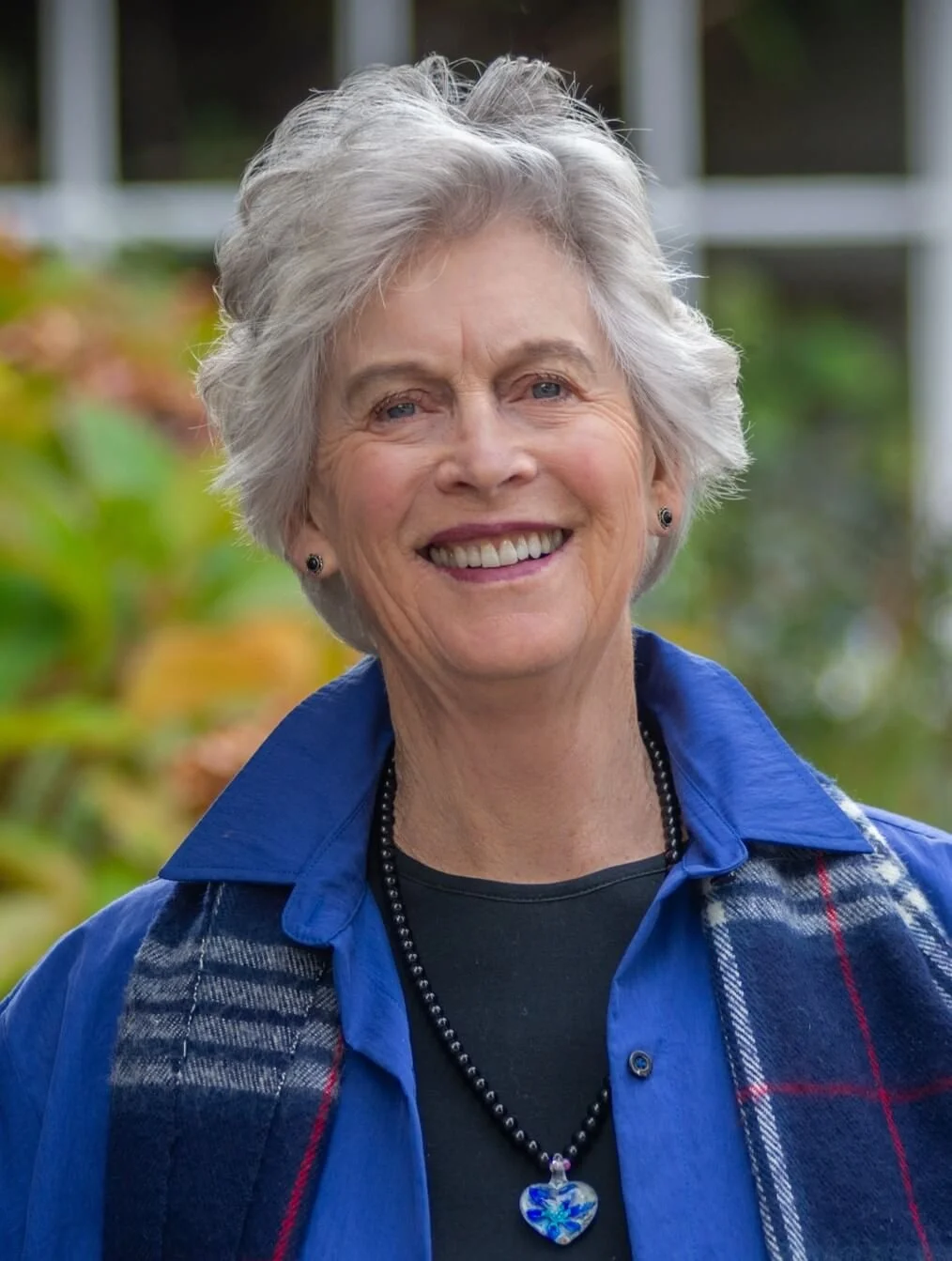 A smiling woman with short gray hair, wearing a blue shirt, a necklace with a heart-shaped pendant, and a plaid scarf, standing outdoors with blurred greenery in the background.