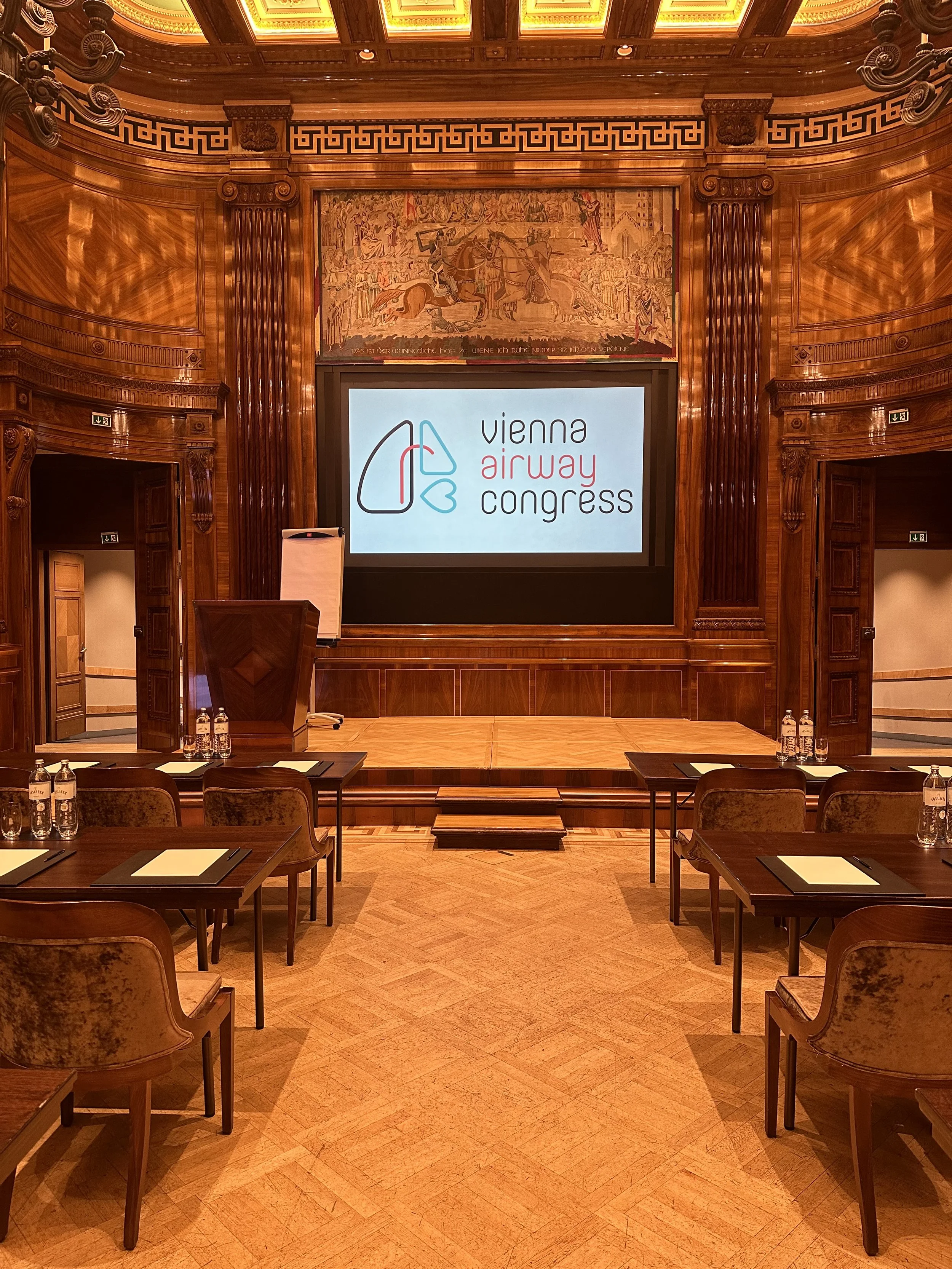 Conference room with wooden walls and ornate decor, set up for a presentation with a large screen displaying the Vienna Airway Congress logo, meeting was to discuss obstructive sleep apnea