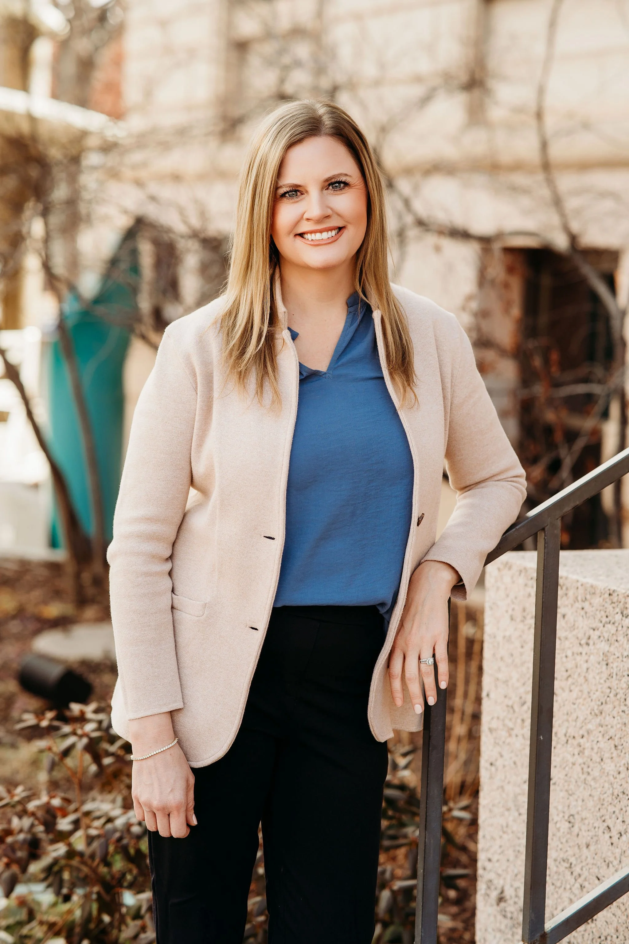 A woman with shoulder-length blonde hair and a beige blazer sitting on a white couch, smiling at the camera, with a green potted plant nearby.