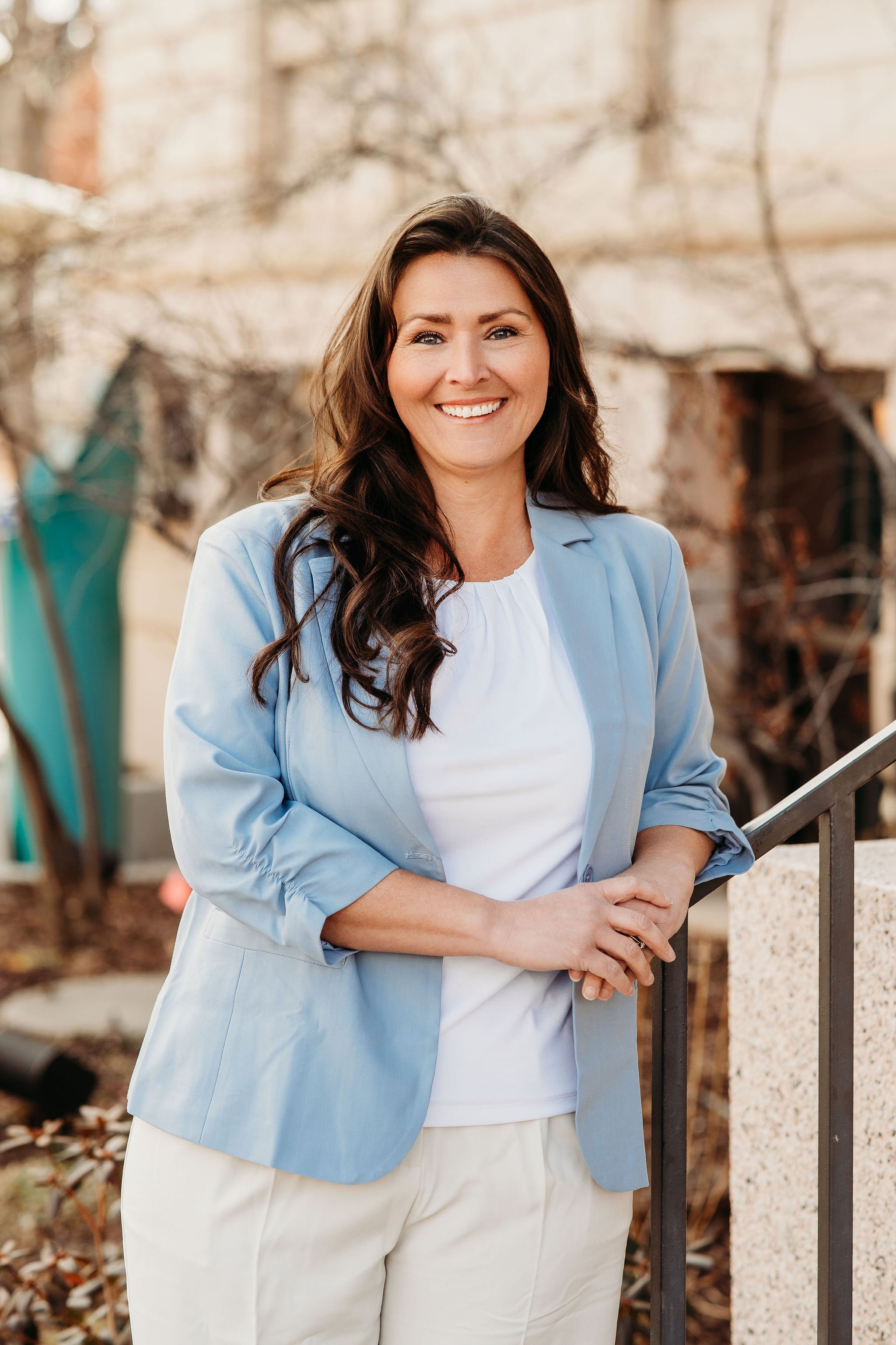 Close-up of a smiling woman with long brown hair and blue eyes, sitting on a brown leather couch against a beige wall.