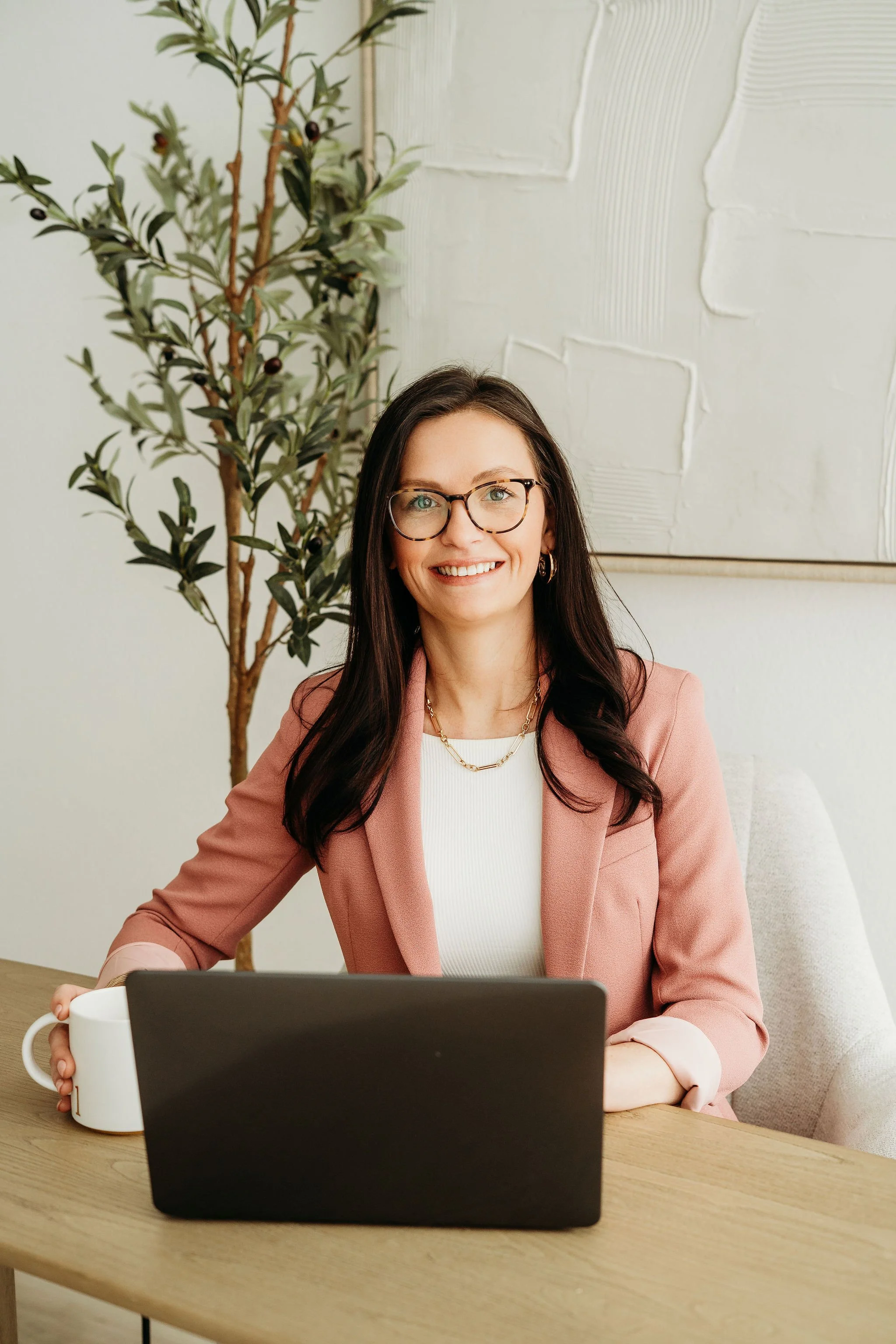A woman with long dark hair, glasses, and a pink blazer smiling at the camera.