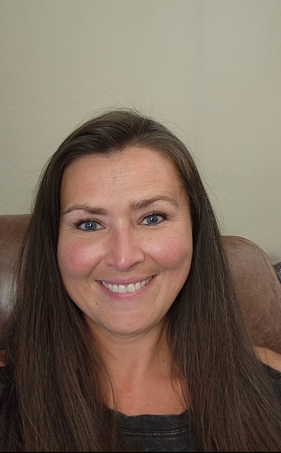 A woman with long brown hair and blue eyes is smiling while sitting on a brown leather chair against a plain light-colored wall.