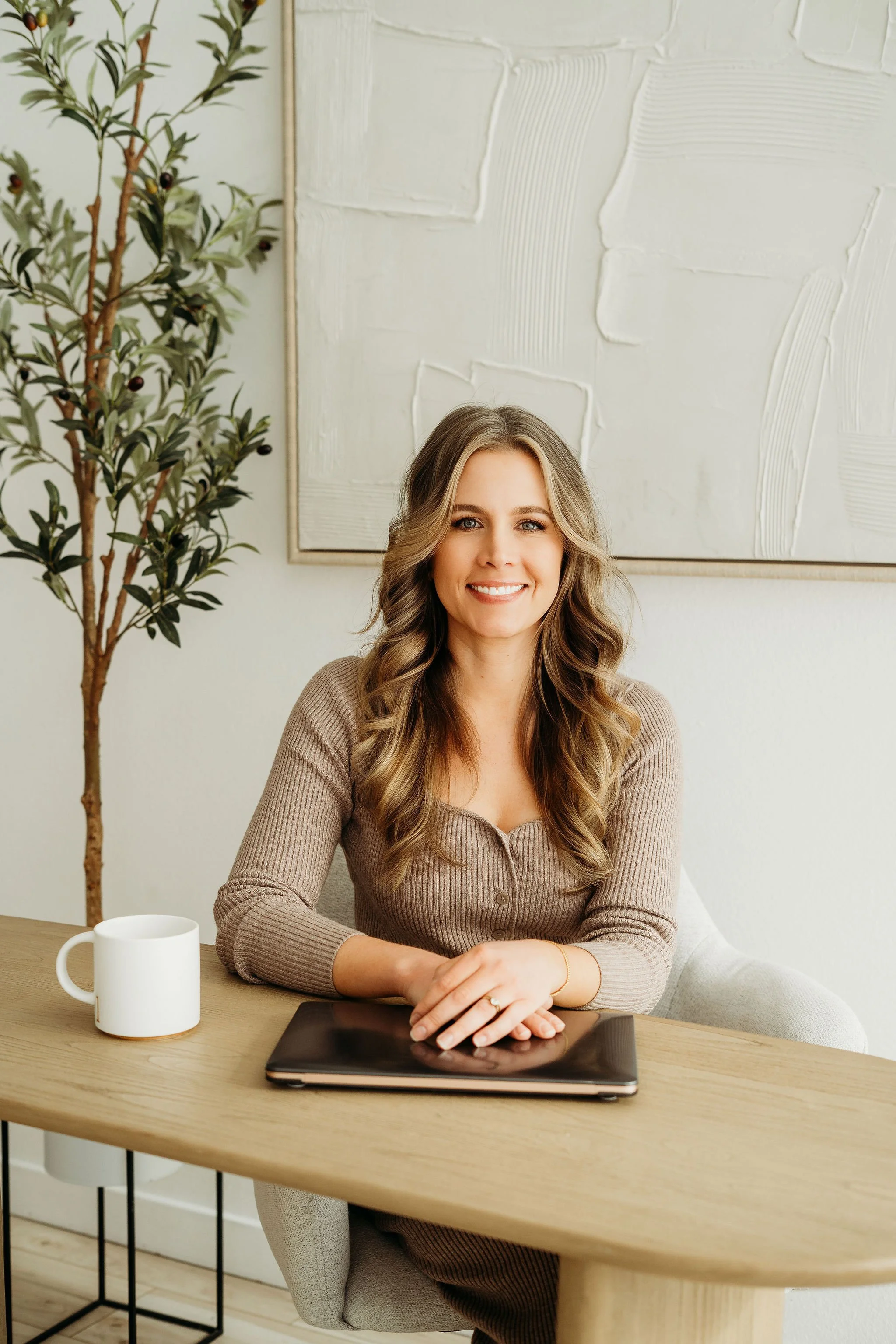A woman with long wavy brown hair and blue eyes, smiling, sitting on a cream-colored armchair in a modern, well-lit office space with a bookshelf and green plant in the background.