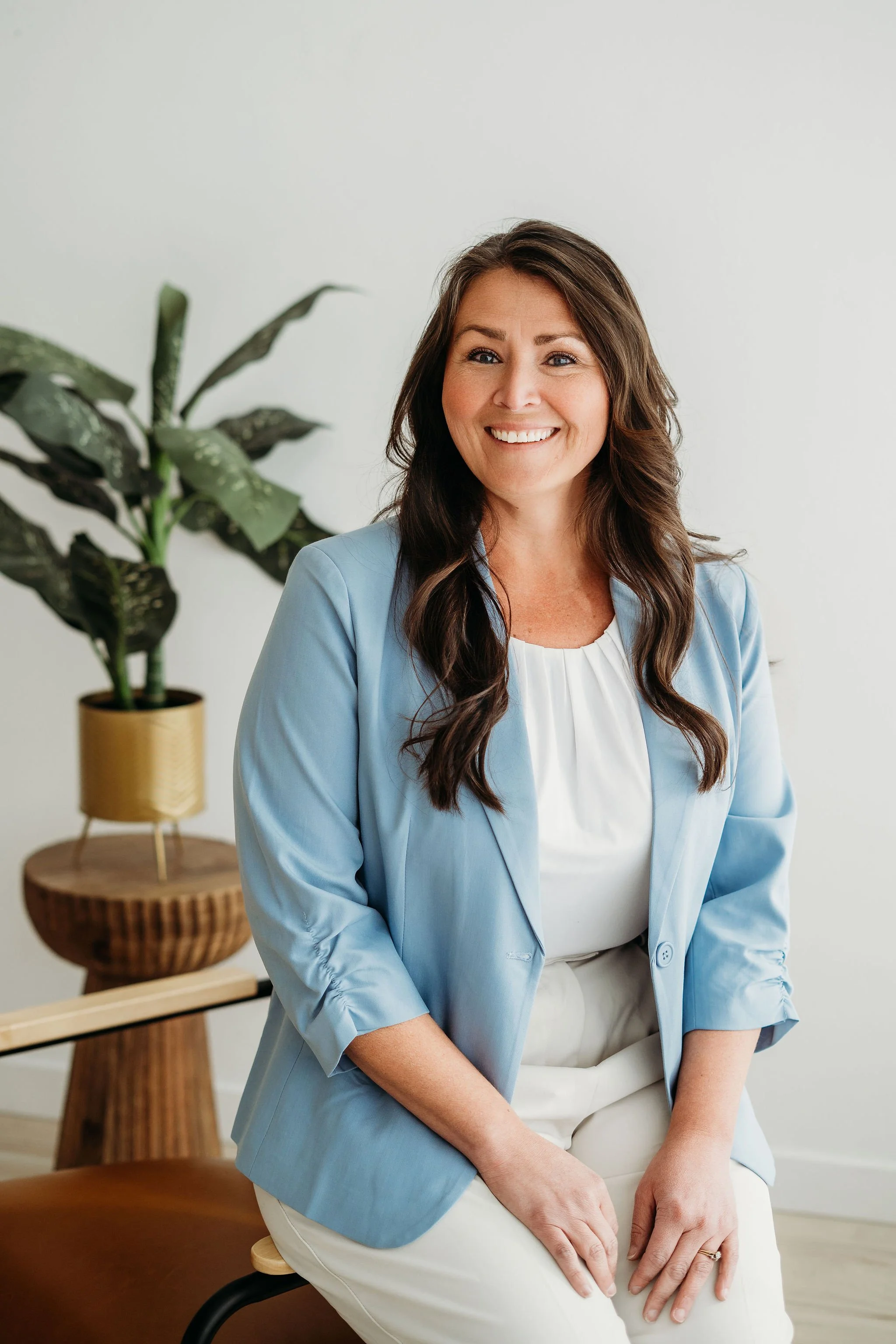 A woman with long dark hair, smiling, wearing a light blue blazer and white blouse, sitting on a chair in a bright, minimalist room with a large leafy plant in the background.
