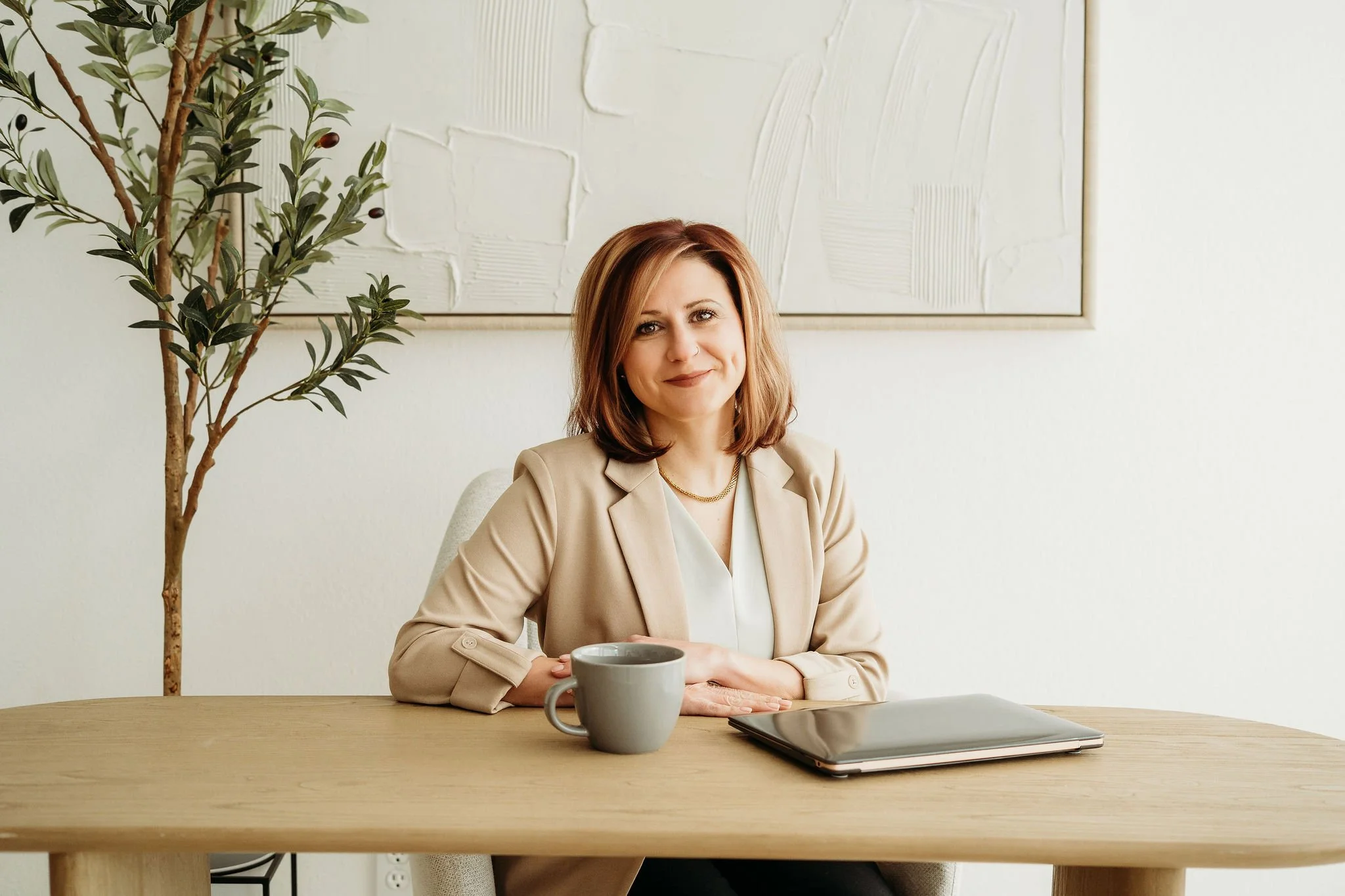 A woman with shoulder-length reddish-brown hair sitting at a wooden desk with a laptop and a gray coffee mug, in a light-colored office with a large abstract textured painting and a potted plant behind her.