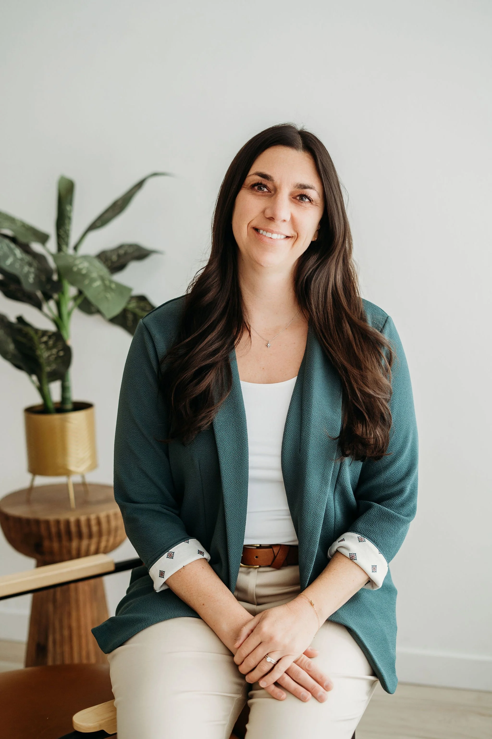 A woman with long brown hair smiling, sitting indoors, wearing a green blazer, white top, and beige pants, with a potted plant on a wooden stand in the background.
