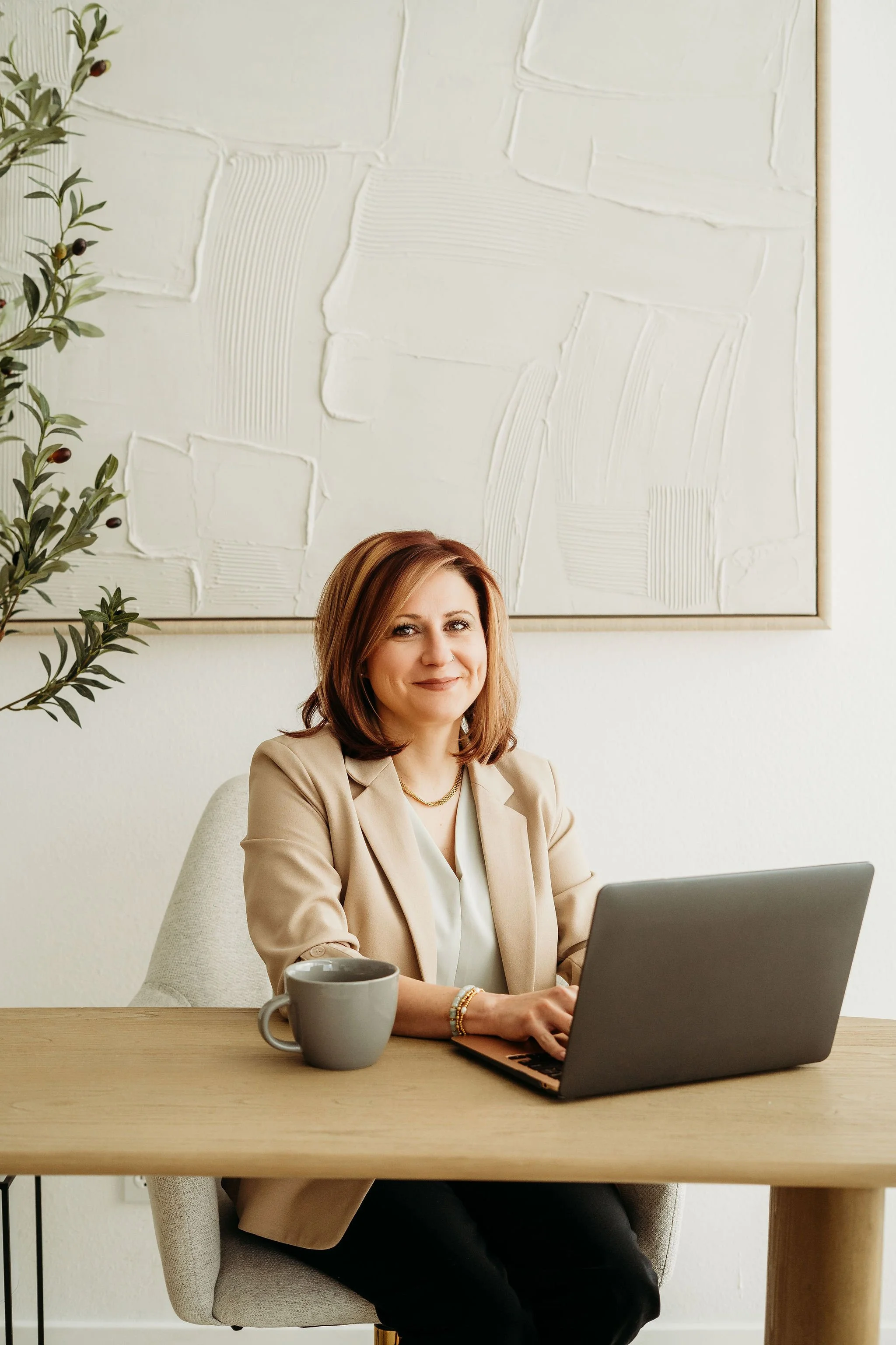 A woman with shoulder-length brunette hair sitting at a light wood desk, working on a laptop. She is wearing a beige blazer and white top, with a coffee mug in front of her and a plant to her left. The background features a white textured wall and a large abstract painting.