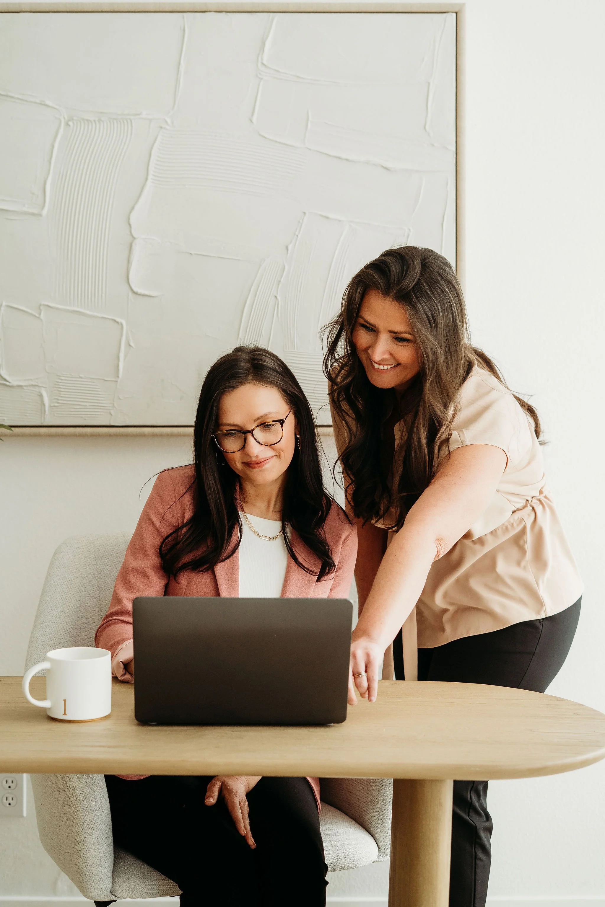 Two women working on a laptop at a desk in an office, one sitting and the other standing and pointing at the screen.