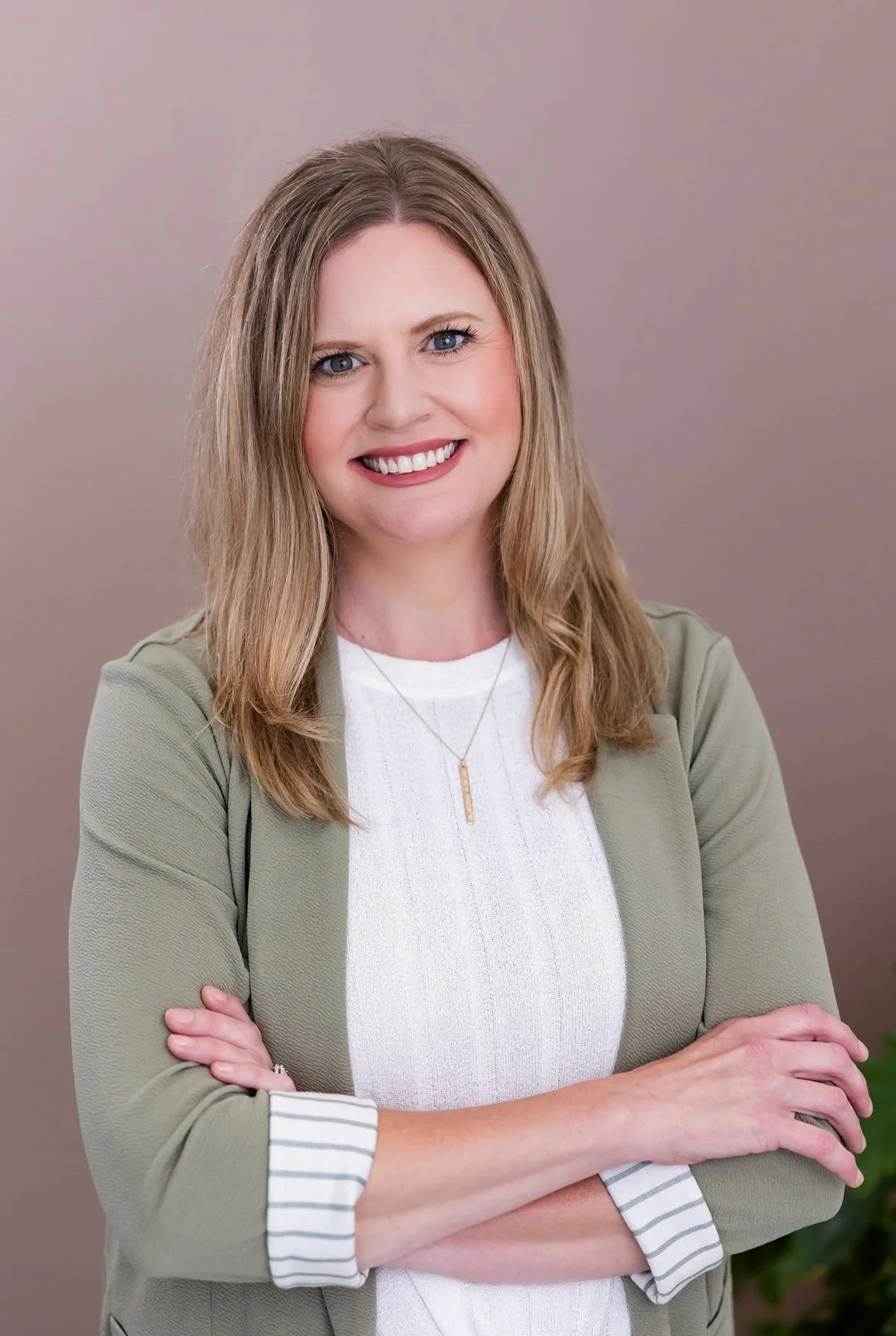 A woman with shoulder-length blonde hair and a beige blazer sitting on a white couch, smiling at the camera, with a green potted plant nearby.