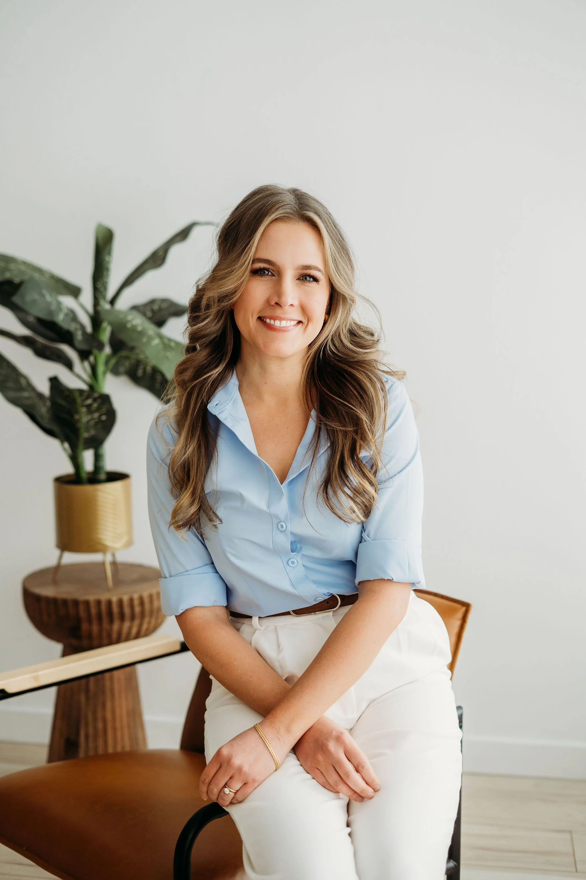 A woman with long, wavy brown hair and blue eyes, wearing a cream blazer and blue jeans, sitting on a wooden bench in a bright, modern room.
