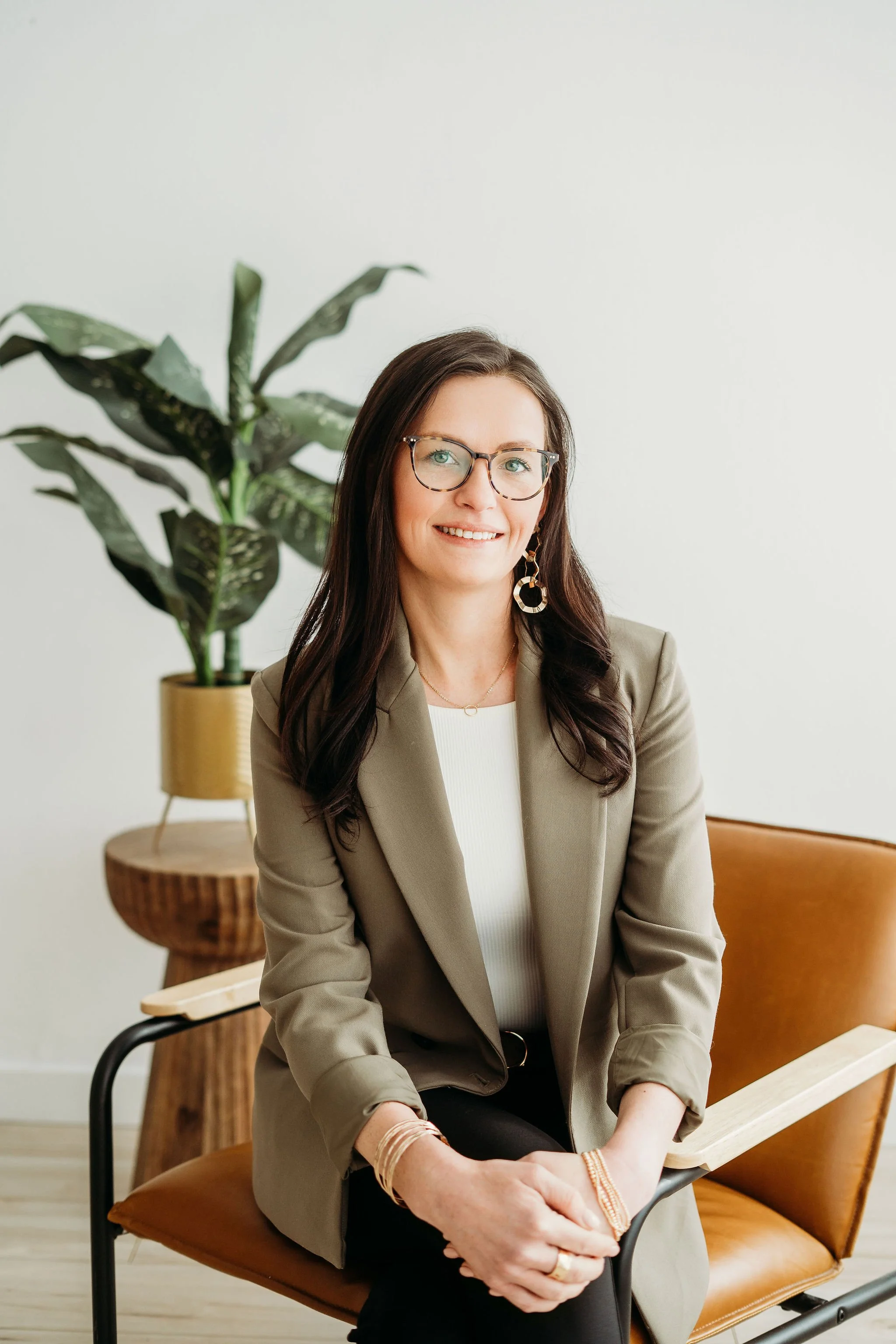 A woman with glasses and long dark hair sitting on a tan leather chair, smiling at the camera, with a large green potted plant and a wooden side table in the background.