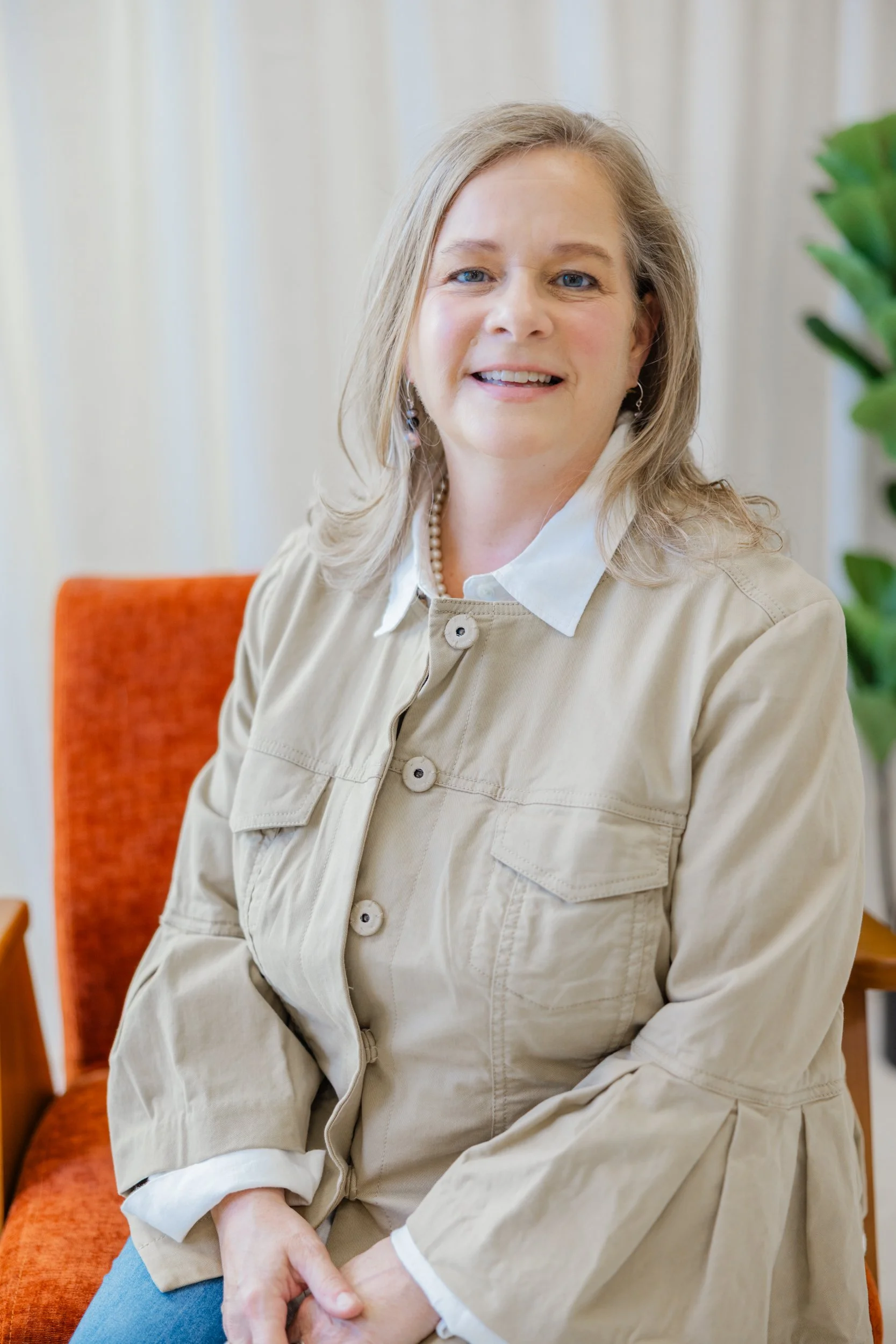 A woman sitting on an orange chair, smiling at the camera, indoors with beige curtains and greenery in the background.
