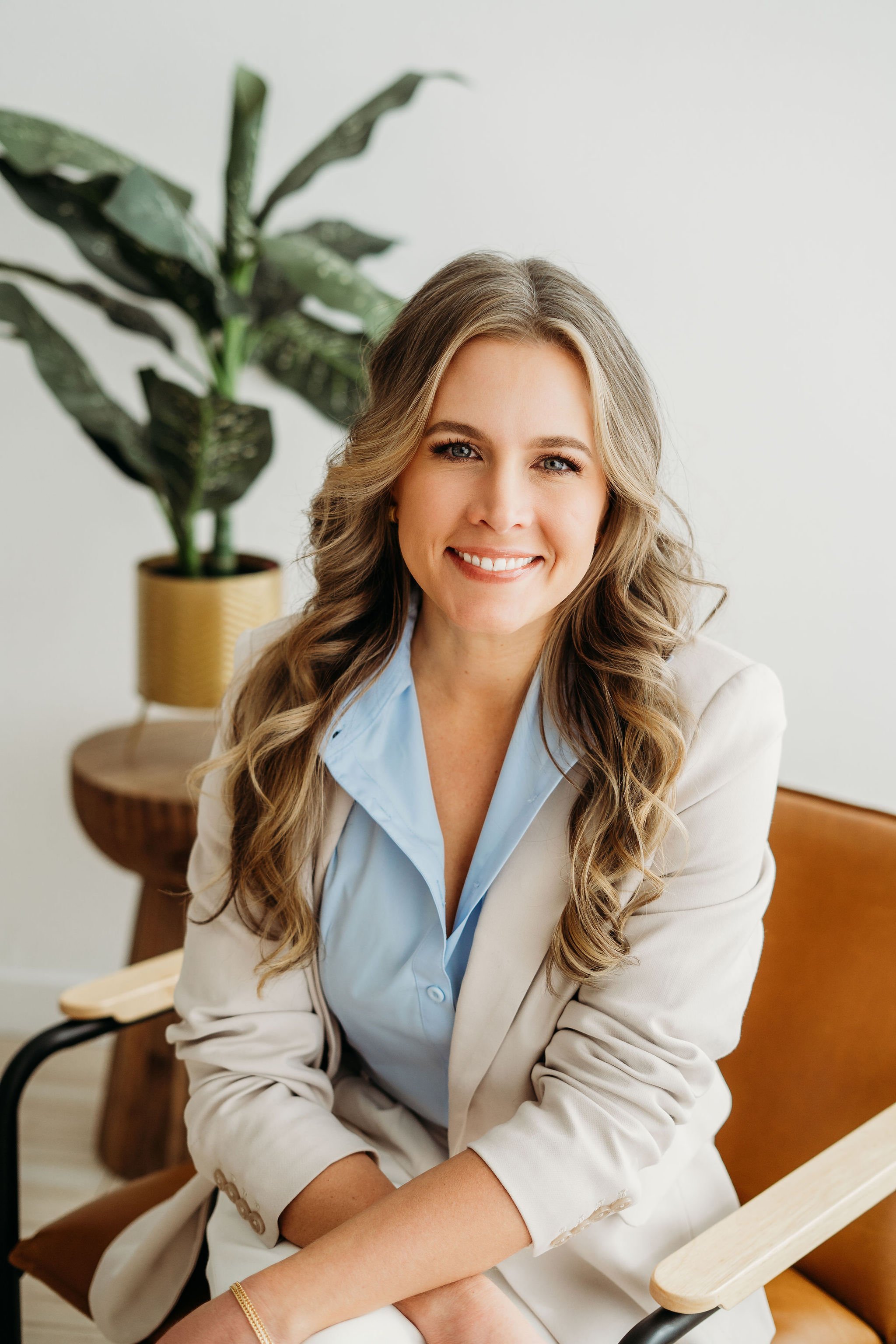 A woman with long, wavy blonde hair, smiling, wearing a light blue shirt and beige blazer, sitting in a room with a large potted plant behind her.