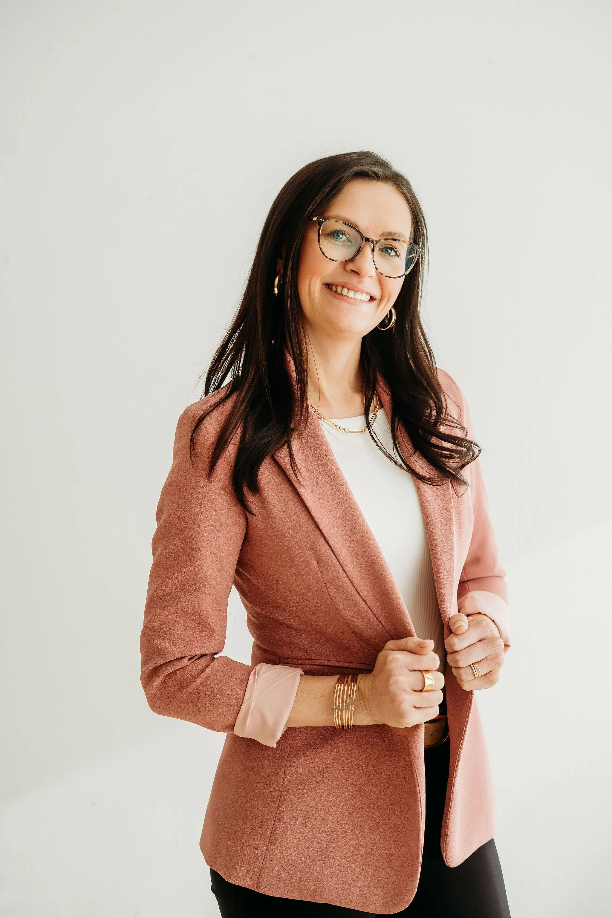 A woman with long dark hair, glasses, wearing a pink blazer and white top, sitting on a cream-colored sofa and smiling.